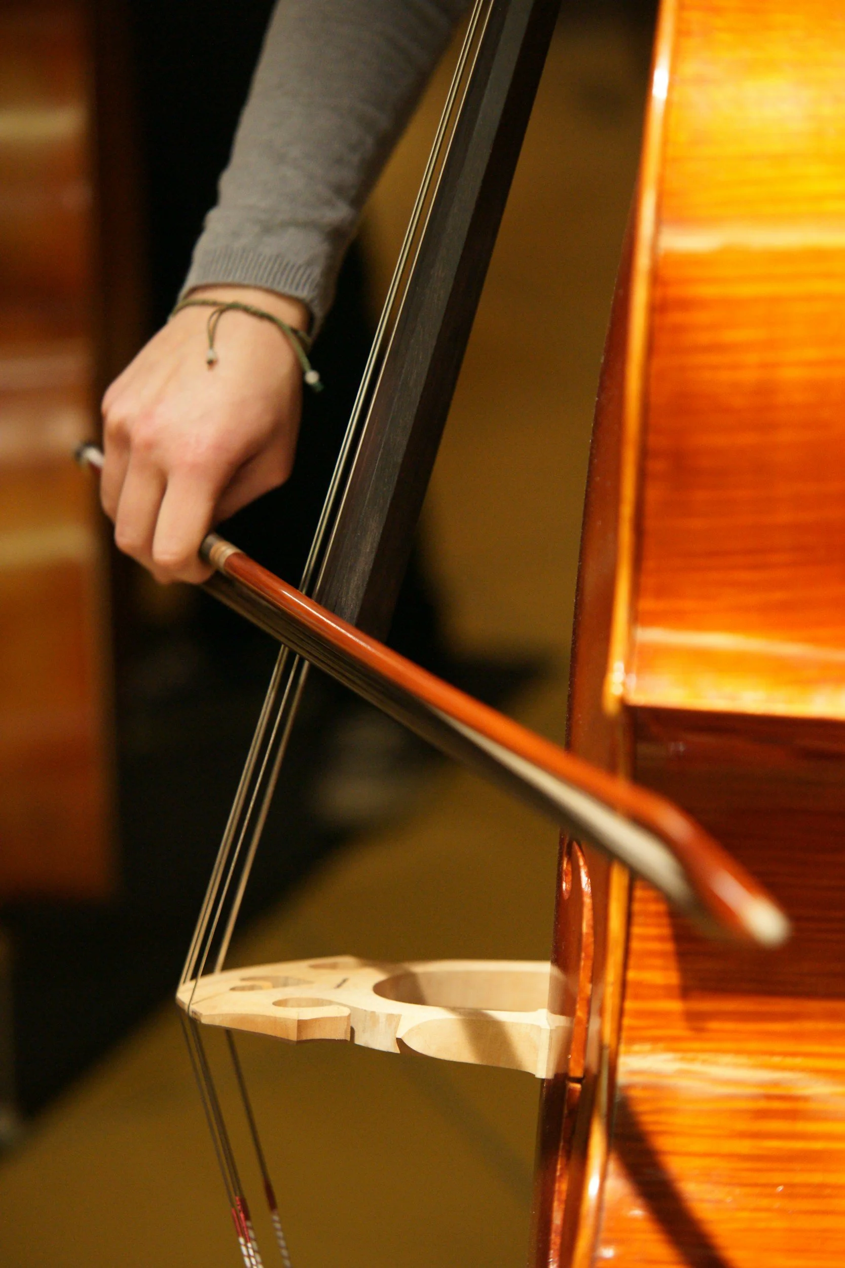 Person's hand holding a violin bow, with a wooden violin and a music stand in the background.