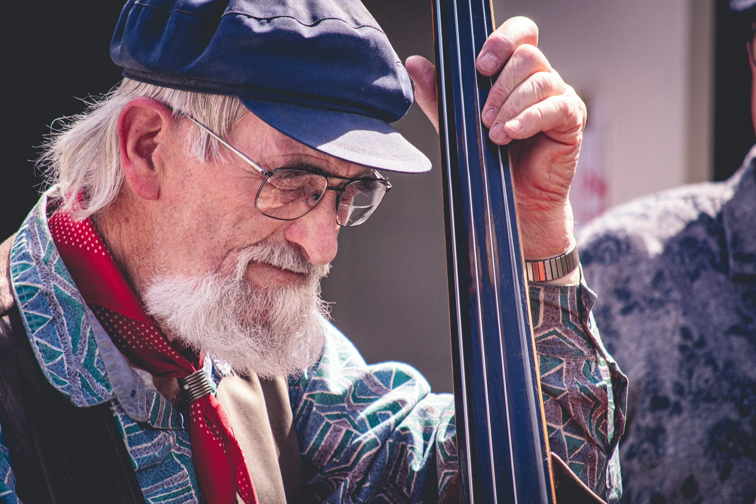 An elderly man with glasses and a white beard playing a double bass. He is wearing a colorful patterned shirt, a red bandana, a blue cap, and a watch. The man appears focused on his music.