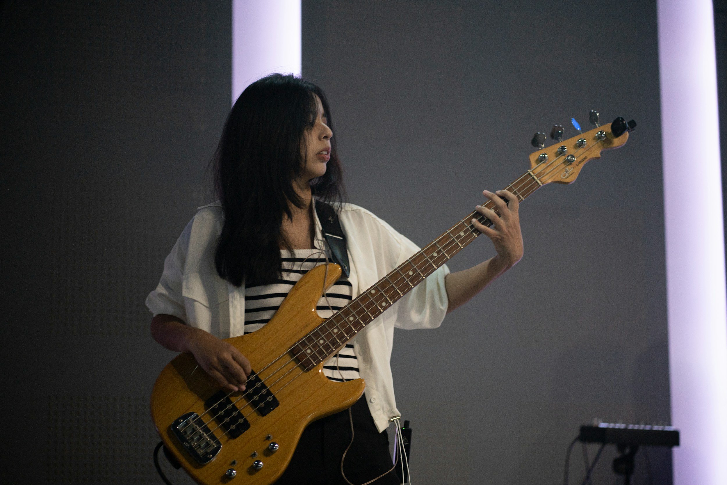 A woman playing a yellow bass guitar in a dimly lit room with purple vertical light strips on the wall.