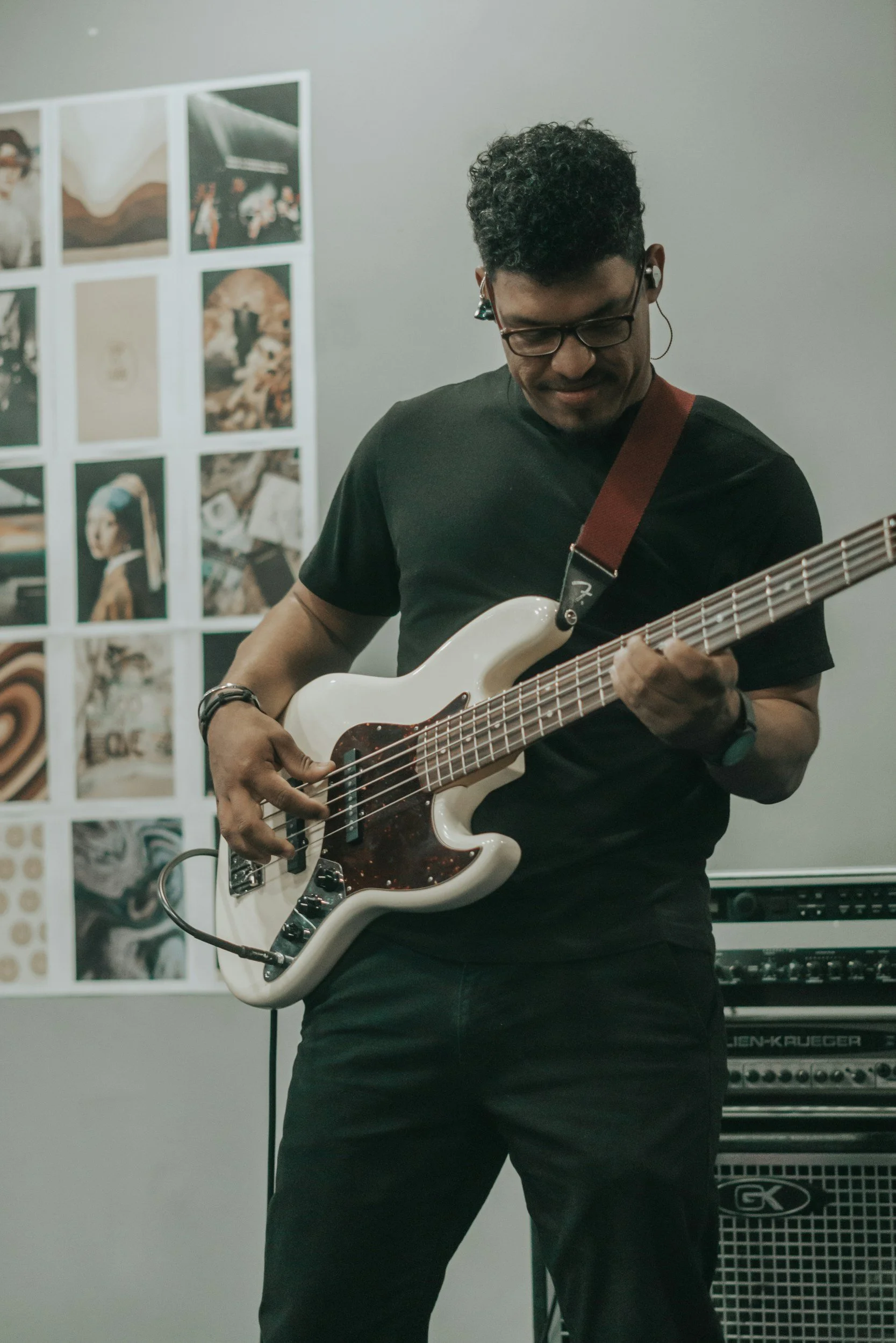 A person playing a white electric guitar in a music studio, with photographs on the wall and equipment in the background.