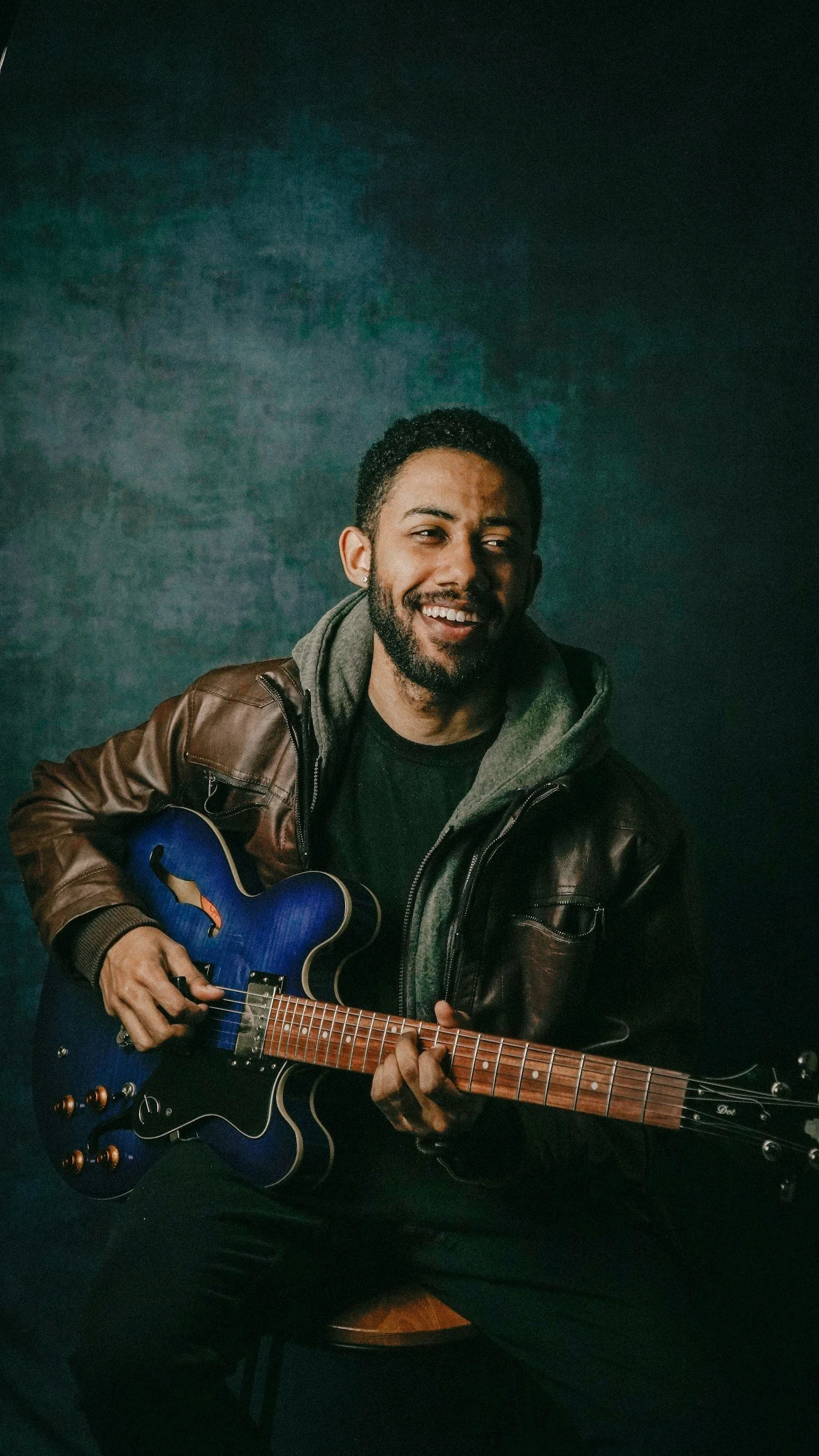 A man with dark curly hair and a beard is smiling and playing a blue acoustic guitar. He is wearing a brown leather jacket layered over a gray hoodie and a black shirt, sitting against a dark green textured background.