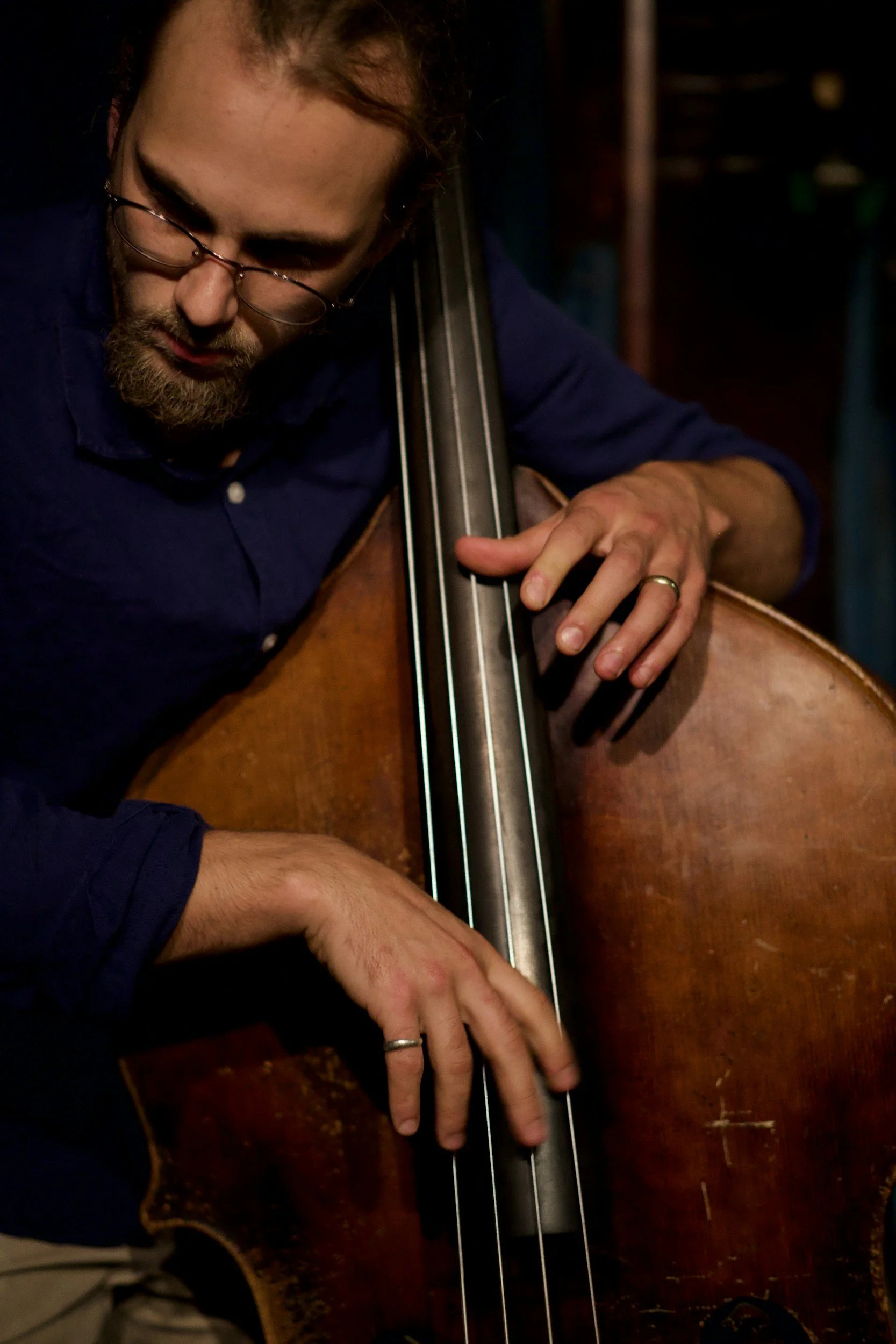 A man with glasses, a beard, and a wedding ring is playing a double bass, focusing intently on his instrument in a dimly lit environment.