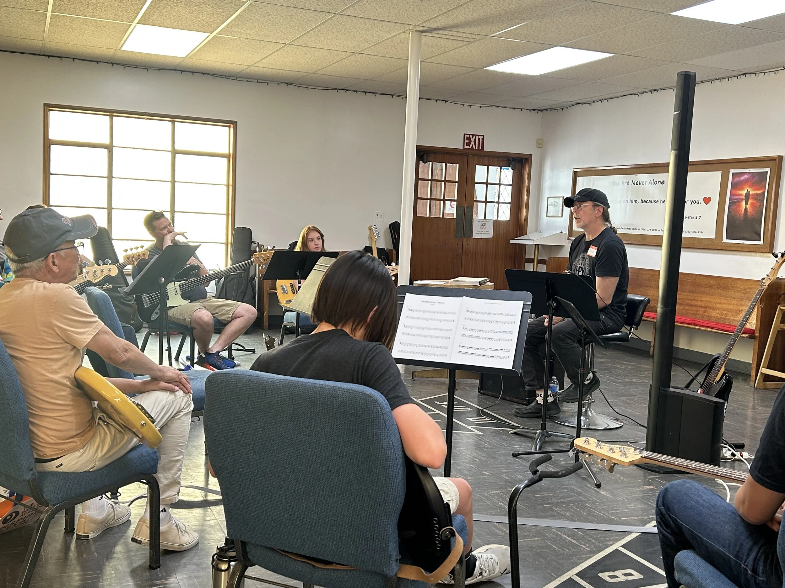 A group of people participating in a music practice or rehearsal session in a room with white walls, wooden trim, and large windows. One person is leading the session, possibly a music teacher or conductor, with sheet music in front of him, while oth