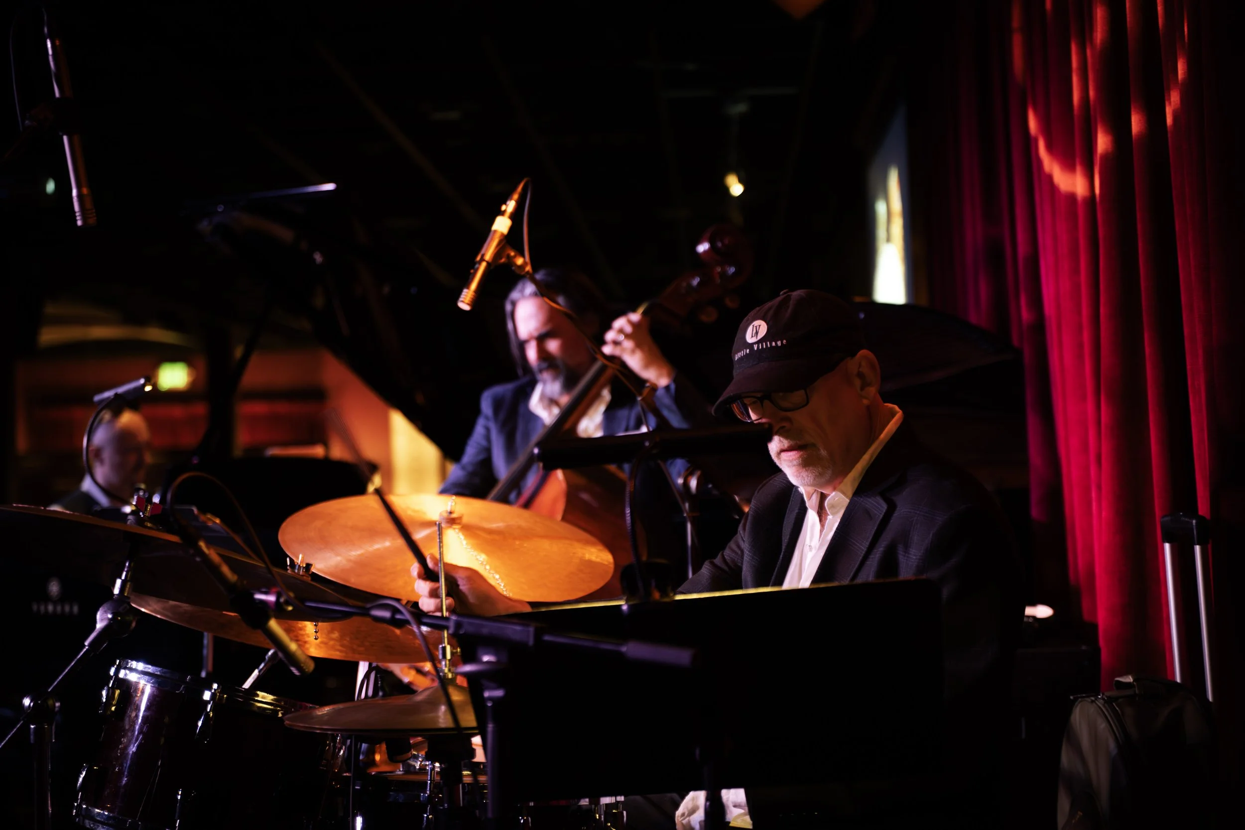 Jazz musicians performing on stage, including a drummer and a double bass player, with red curtains in the background.