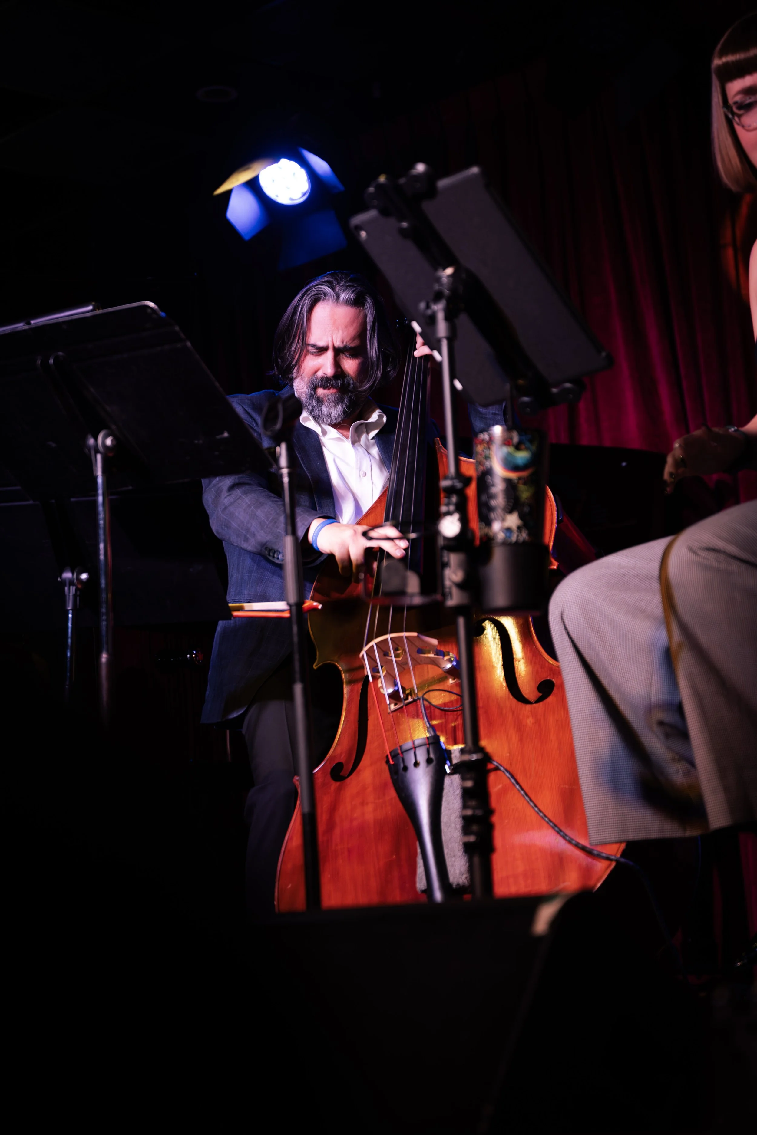 A man with long dark hair and a beard playing a cello on stage, with music stands and stage lighting visible.