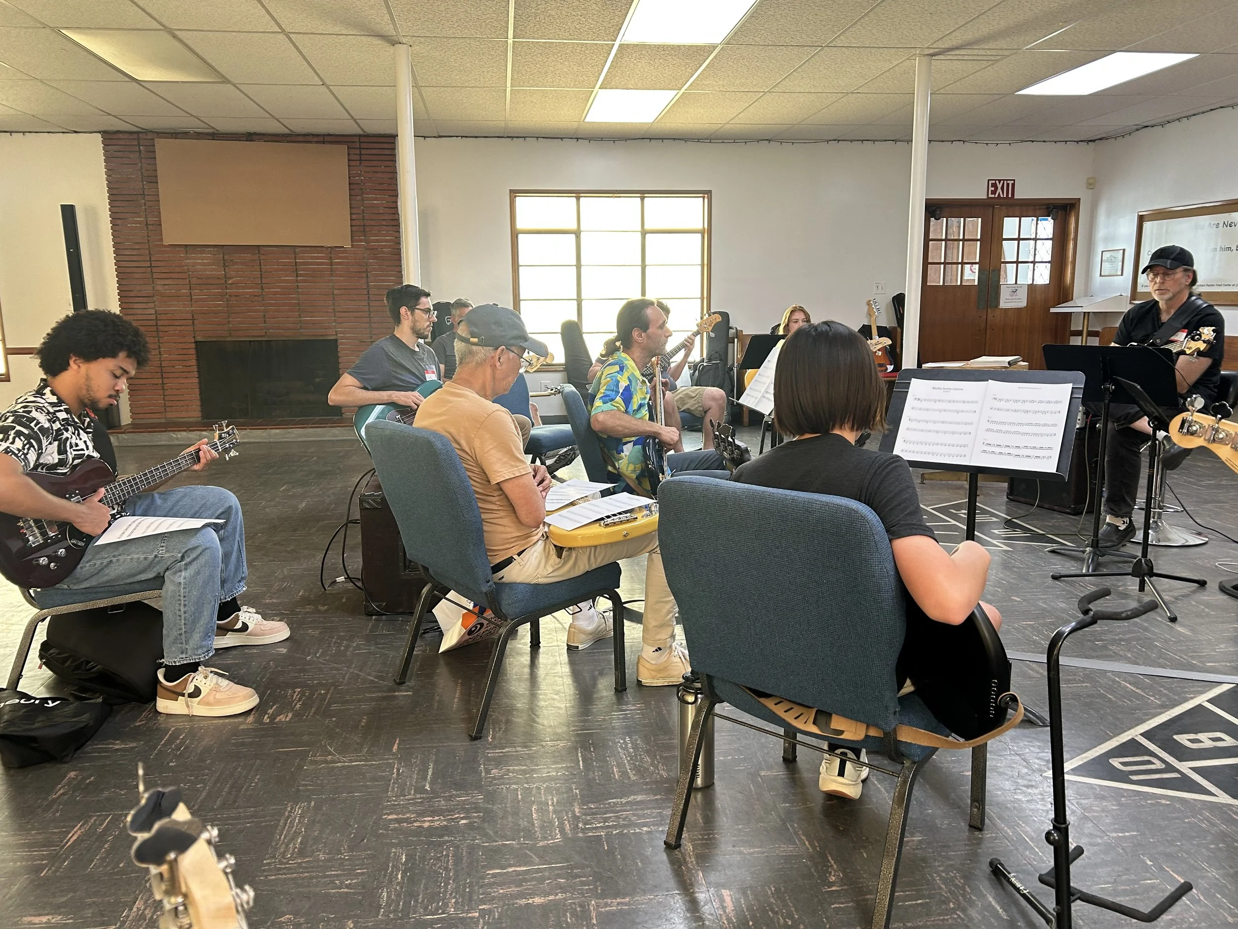 A group of people sitting in a room playing guitars and reading sheet music, with one person standing and playing an electric guitar.