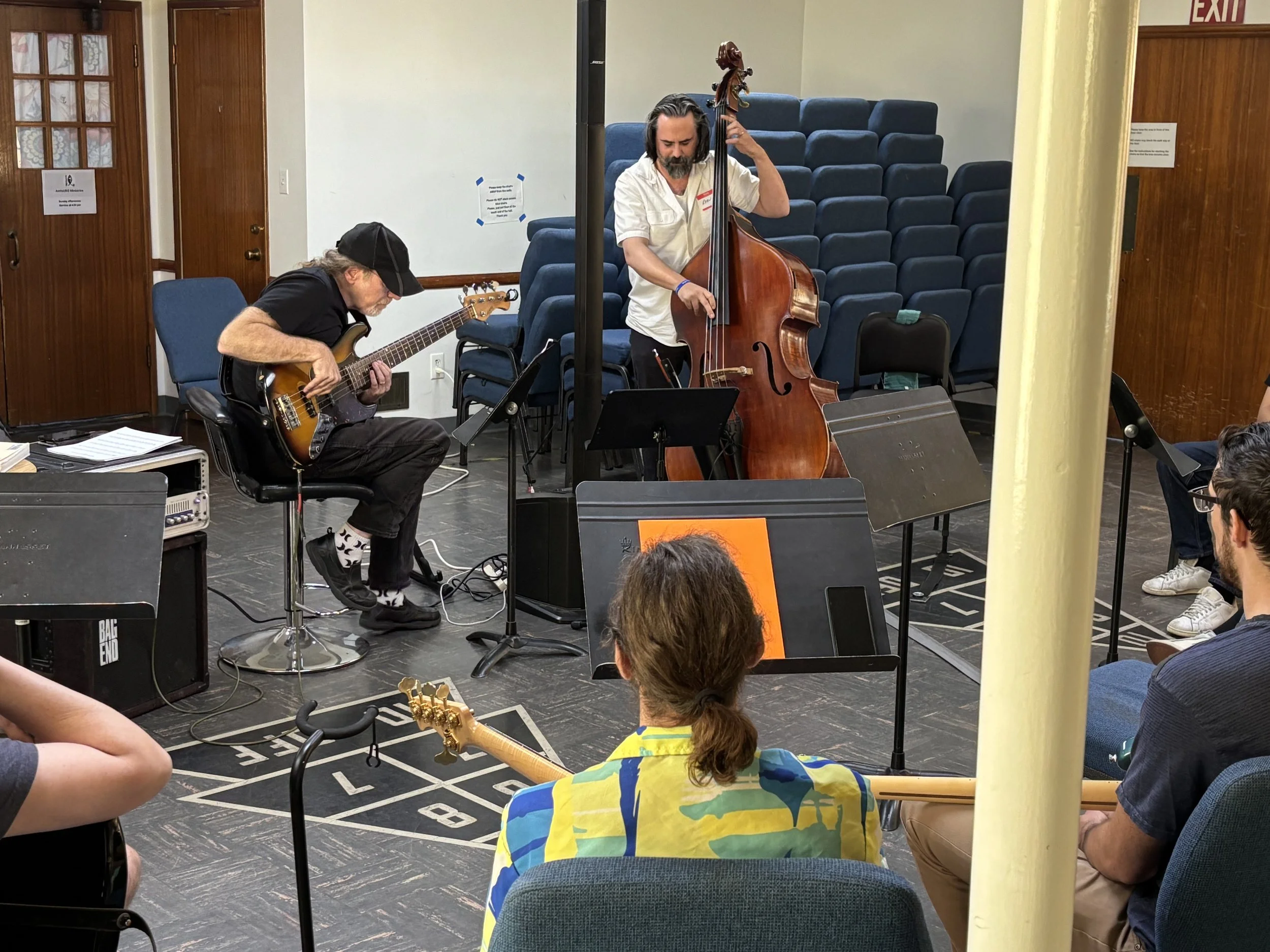 Musicians performing in a room with blue chairs stacked on the walls, audience members sitting and watching, and a wooden door with a sign on it.