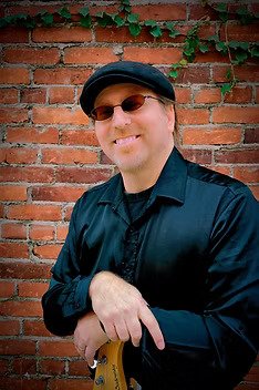 A smiling man wearing sunglasses, a black hat, and a black jacket, pointing his finger downward in front of a brick wall with greenery.