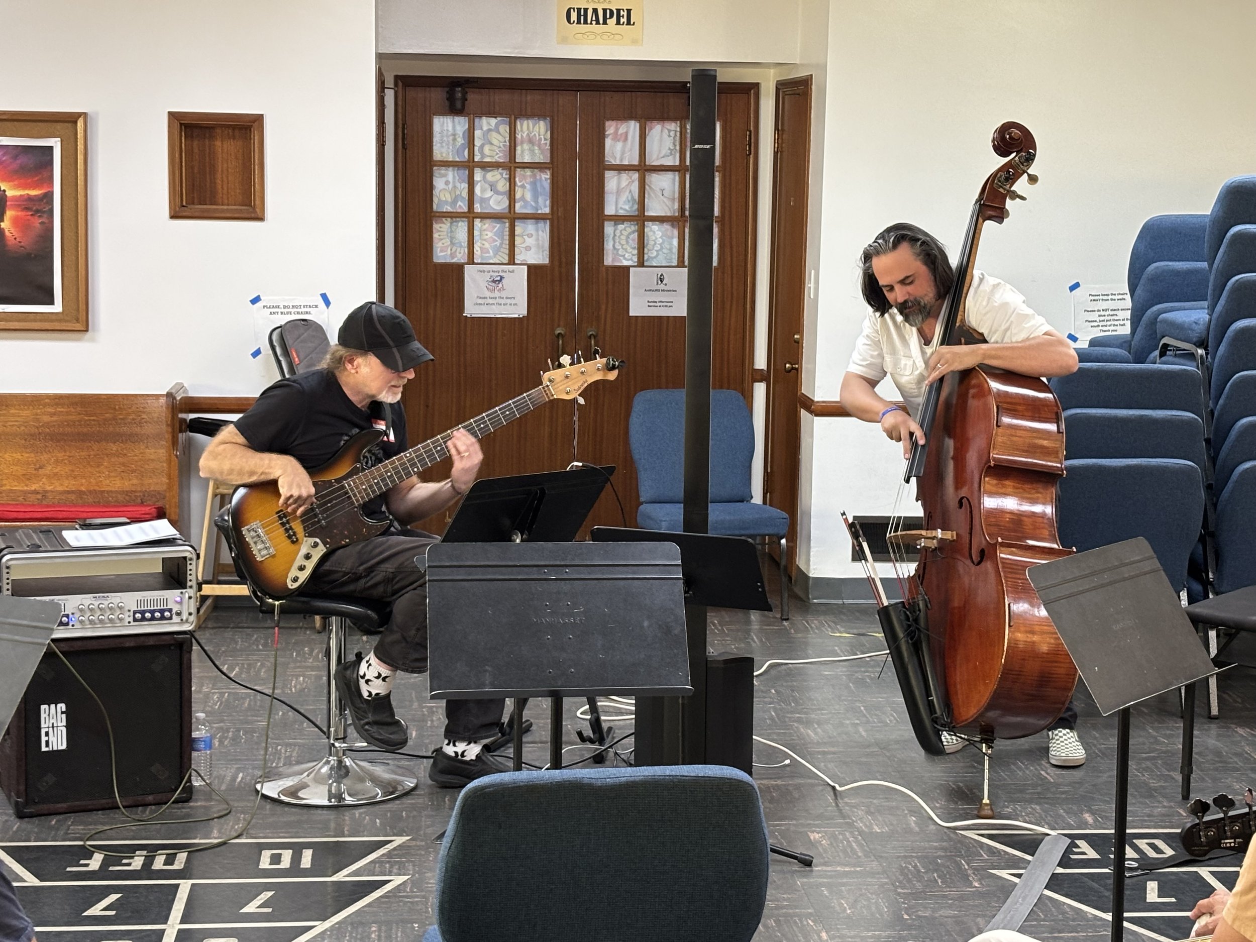 Two musicians playing string instruments in a community hall, with blue chairs and signs on the doors.