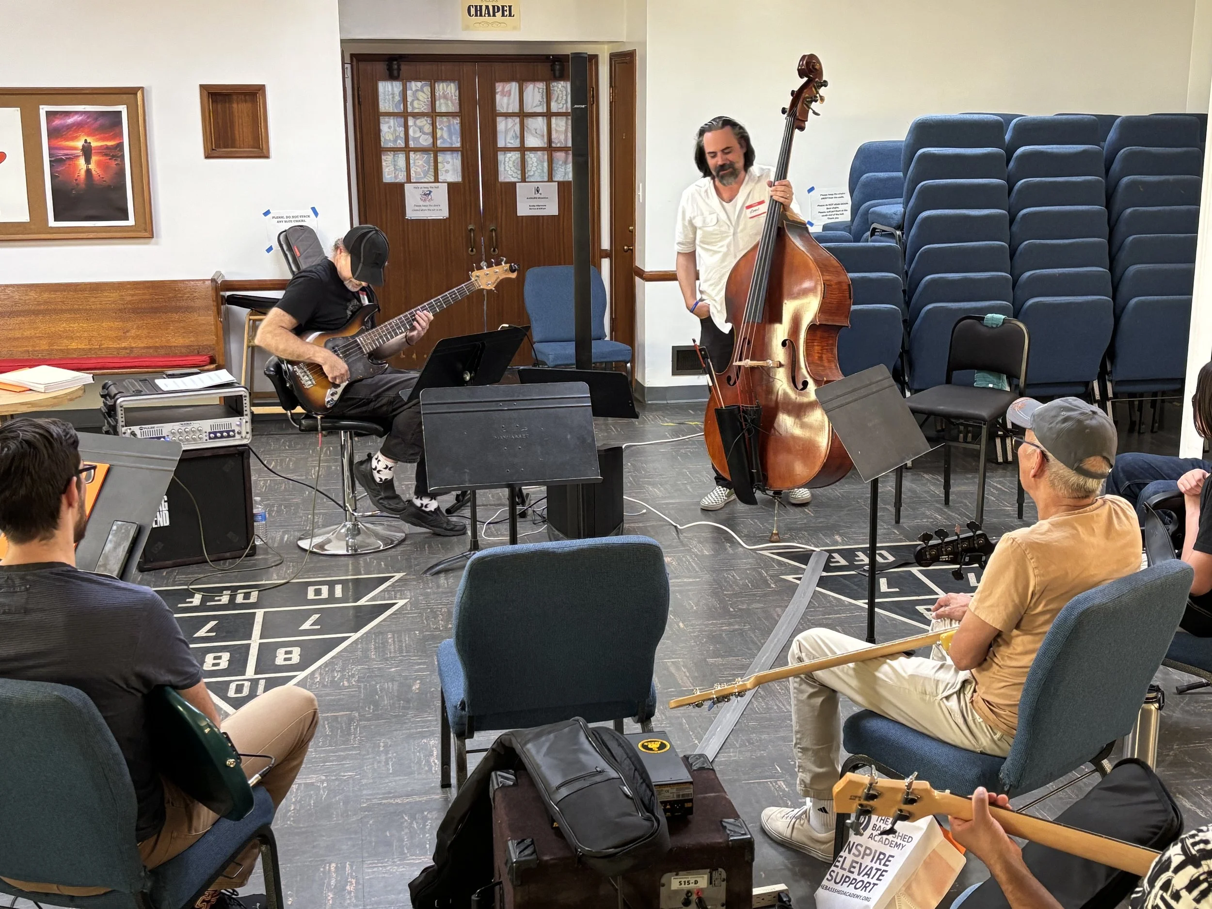 Two musicians playing instruments in a small indoor venue, one on bass and the other on guitar, with seated audience members watching.