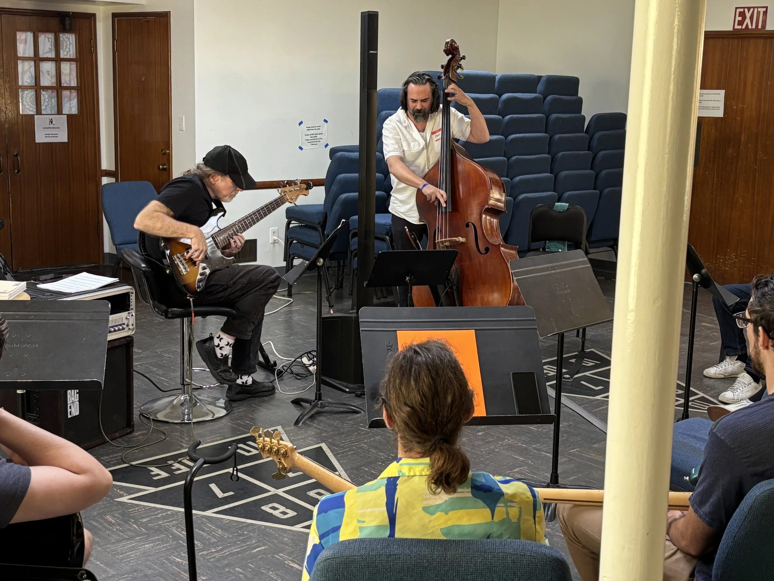 Two musicians perform in a room filled with seated audience members. One plays an electric guitar, and the other plays a double bass. The audience members are sitting on chairs and are watching the performance. The room has wooden doors, and there ar