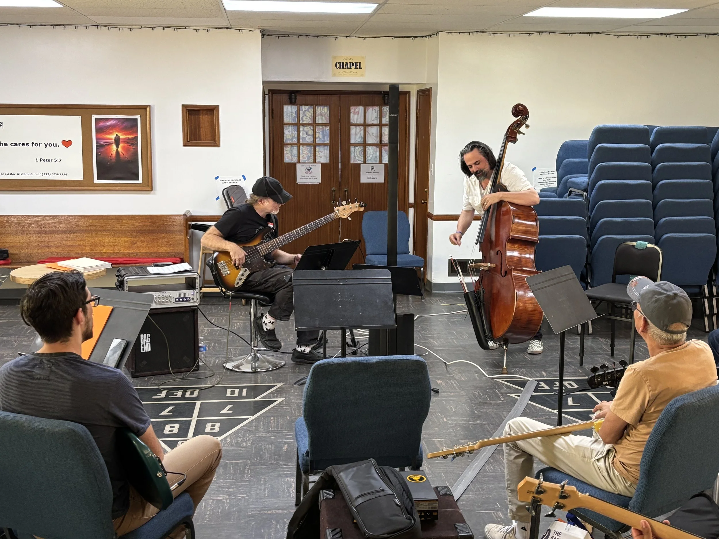 Two musicians, one playing an electric guitar and the other playing a double bass, perform in front of an audience in a room with chairs and a door labeled 'Chapel.'