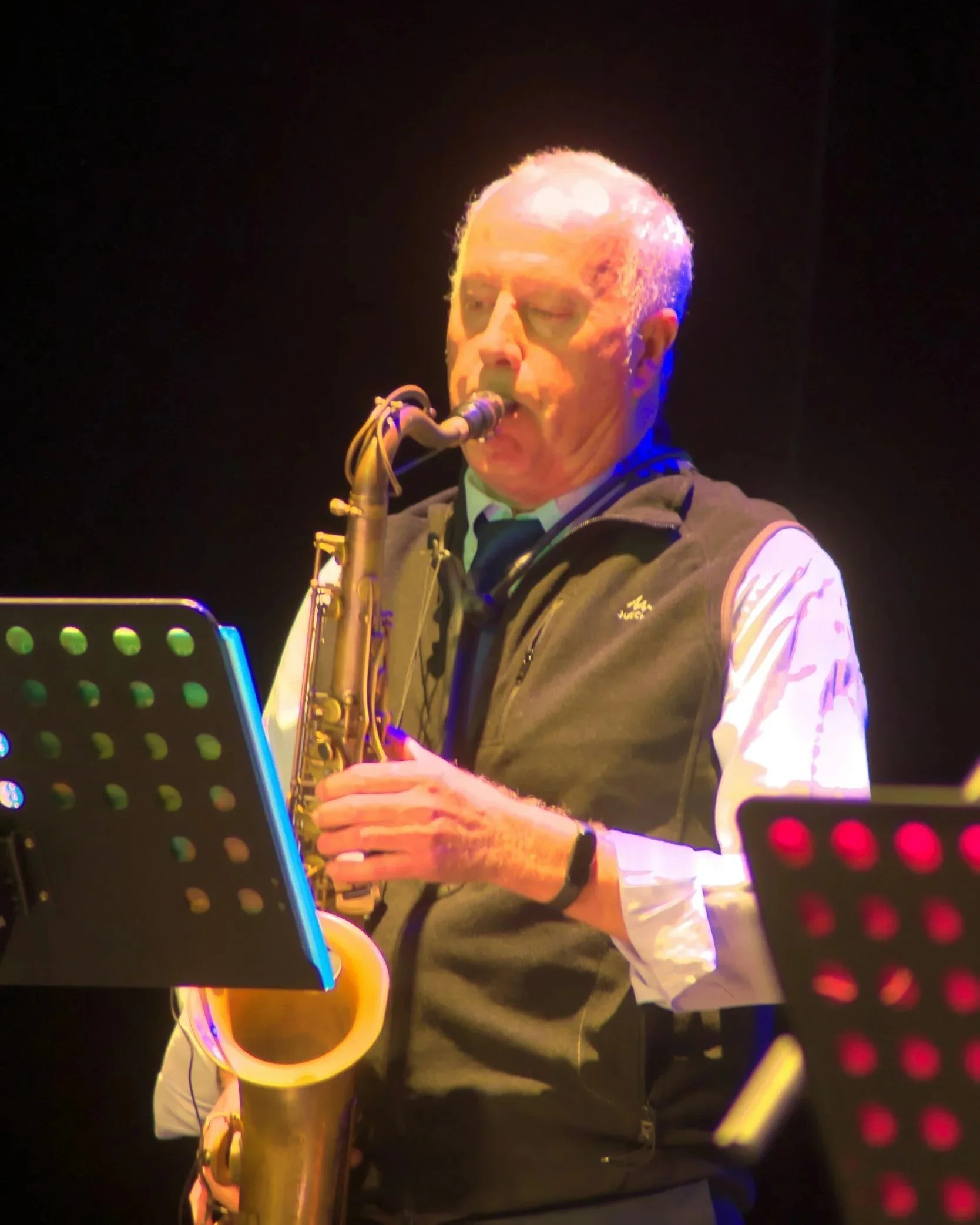 A male musician playing a saxophone on stage, with stage lights reflecting off his face and instrument.