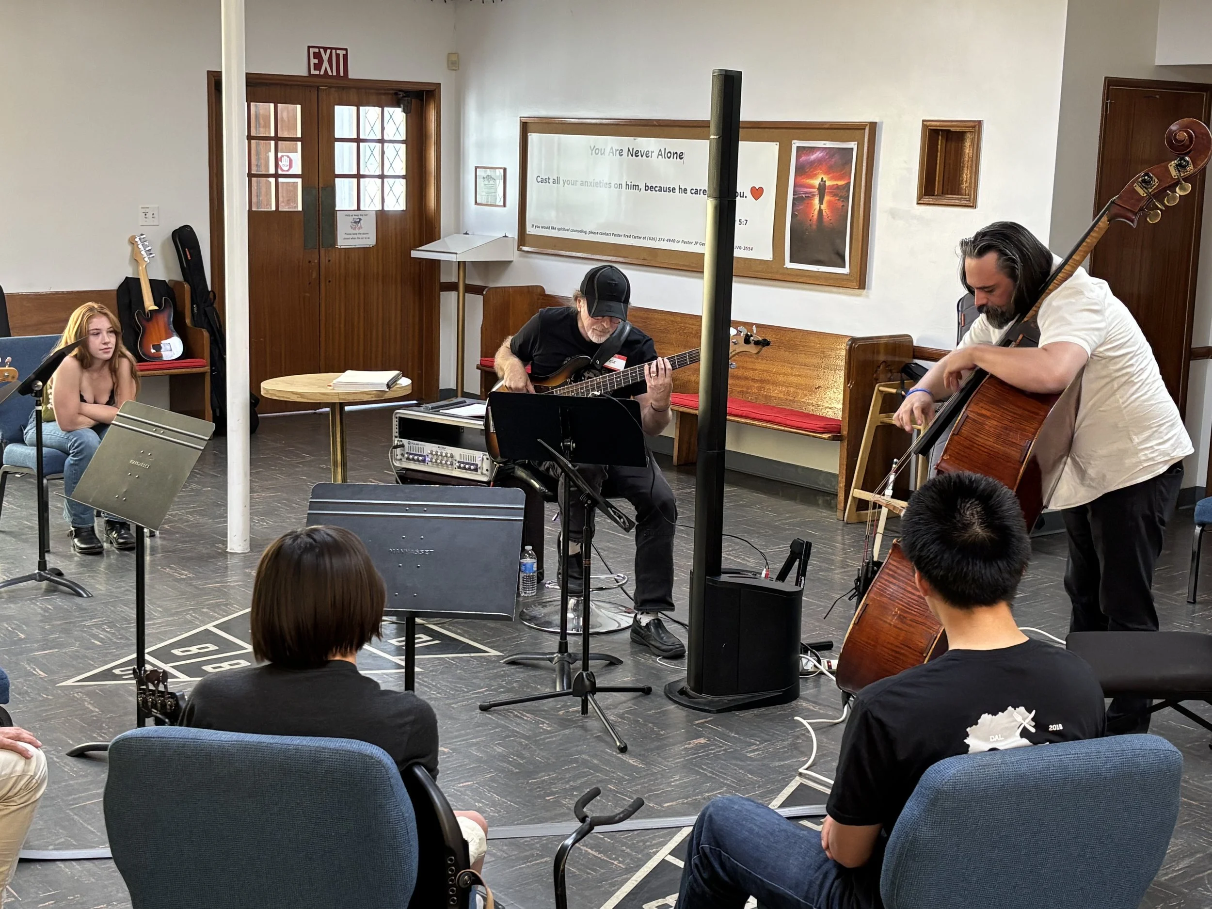 A small indoor concert with three musicians playing string instruments and an audience seated in chairs. One musician is playing a guitar, another a double bass, and a third a bass guitar. The room has posters and a sign on the wall, with an exit doo