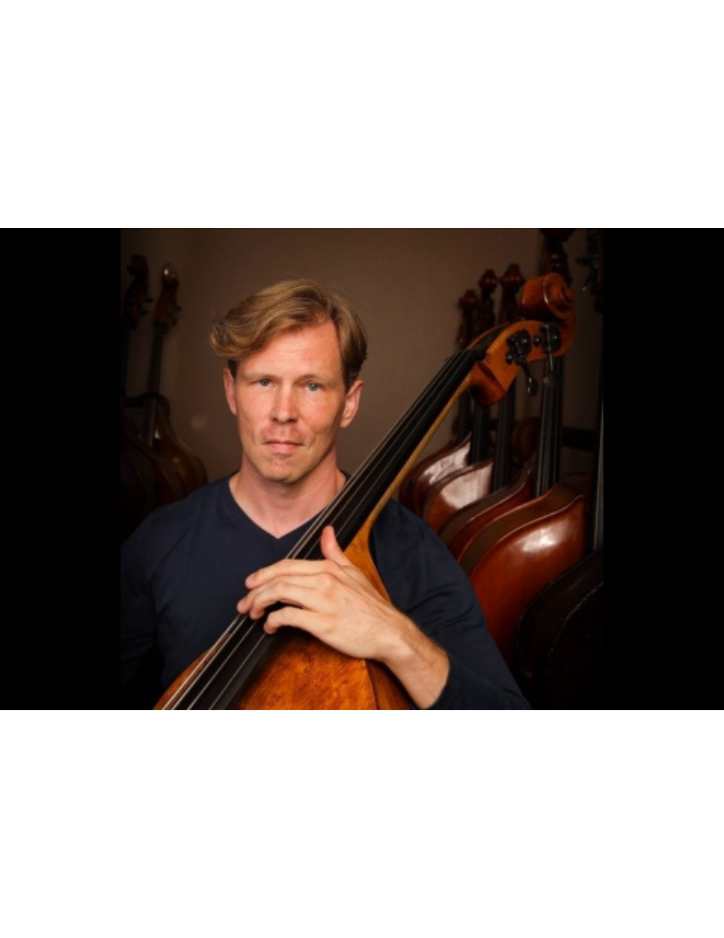 Portrait of a man holding a double bass with a background of several other string instruments.