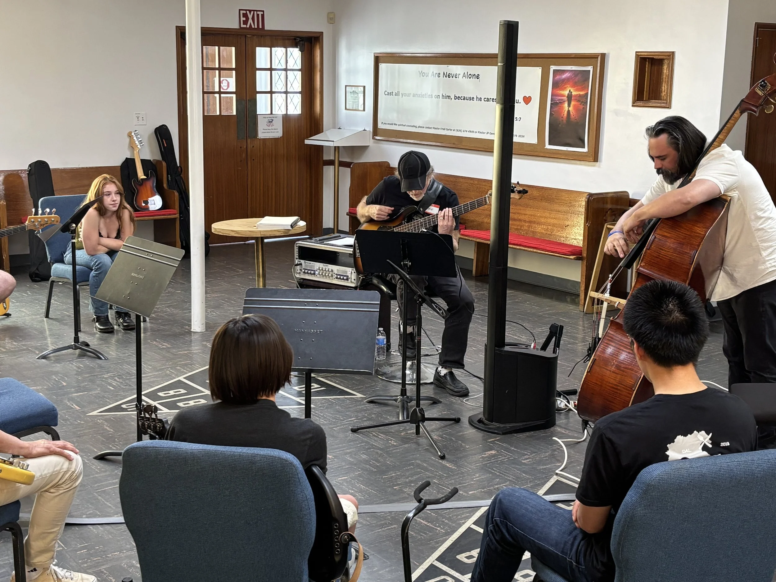 A group of musicians performing in a room with wooden paneling and a sign on the wall that reads "You Are Never Alone." The musicians are playing various string instruments including a guitar and a double bass, with audience members seated nearby.