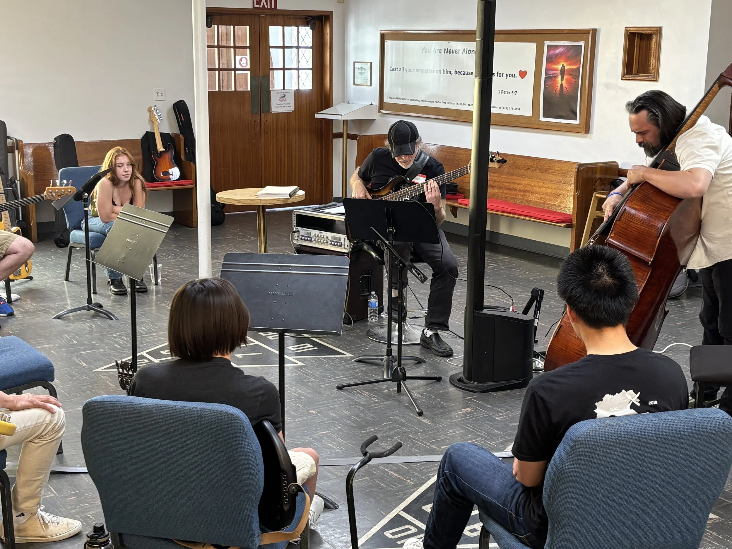 A group of musicians rehearsing in a small room with audience members seated in chairs. The musicians include a woman with long red hair, a man with long dark hair, and an older man with a beard, all playing guitars. There is a drum set and music sta