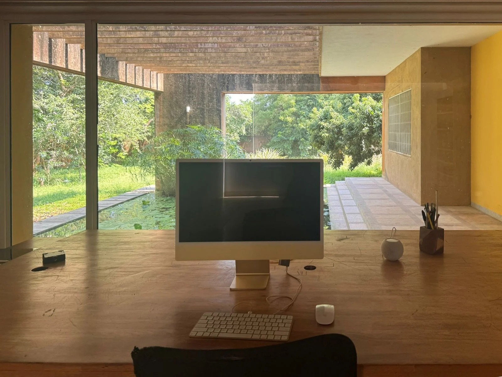 Office workspace with an iMac computer, keyboard, and mouse on a wooden desk, overlooking a lush green garden through large glass windows.
