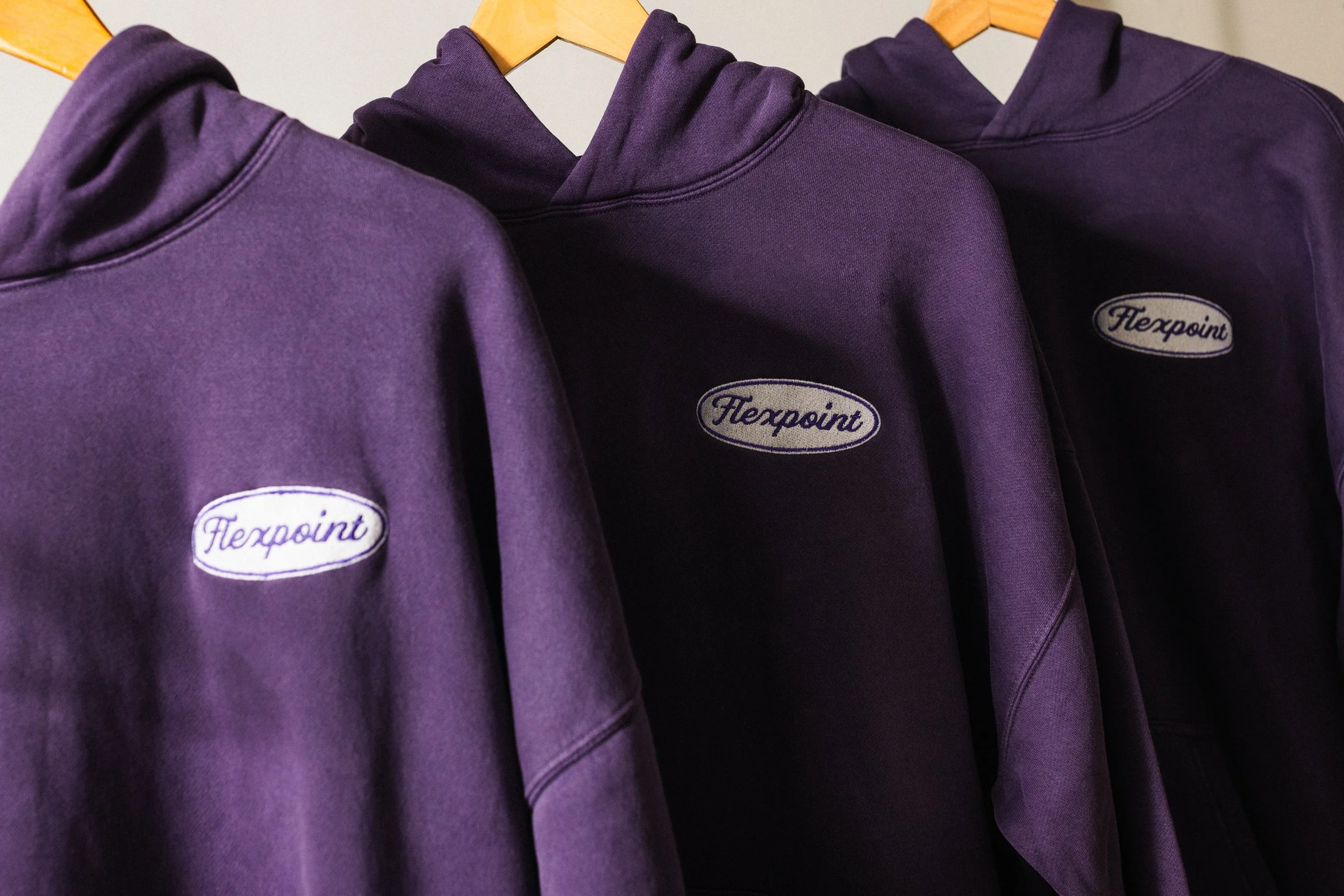 Three purple hoodies hanging on wooden hangers, each with a small oval logo patch that says 'Flexpoint.'