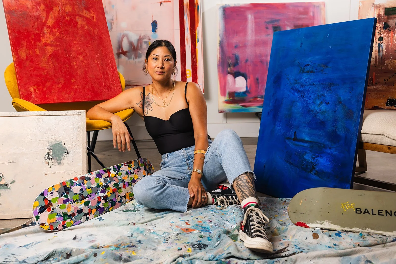 A woman with tattoos wearing a black top and jeans, sitting on the floor among colorful abstract paintings, skateboards, and a painted canvas in an art studio.
