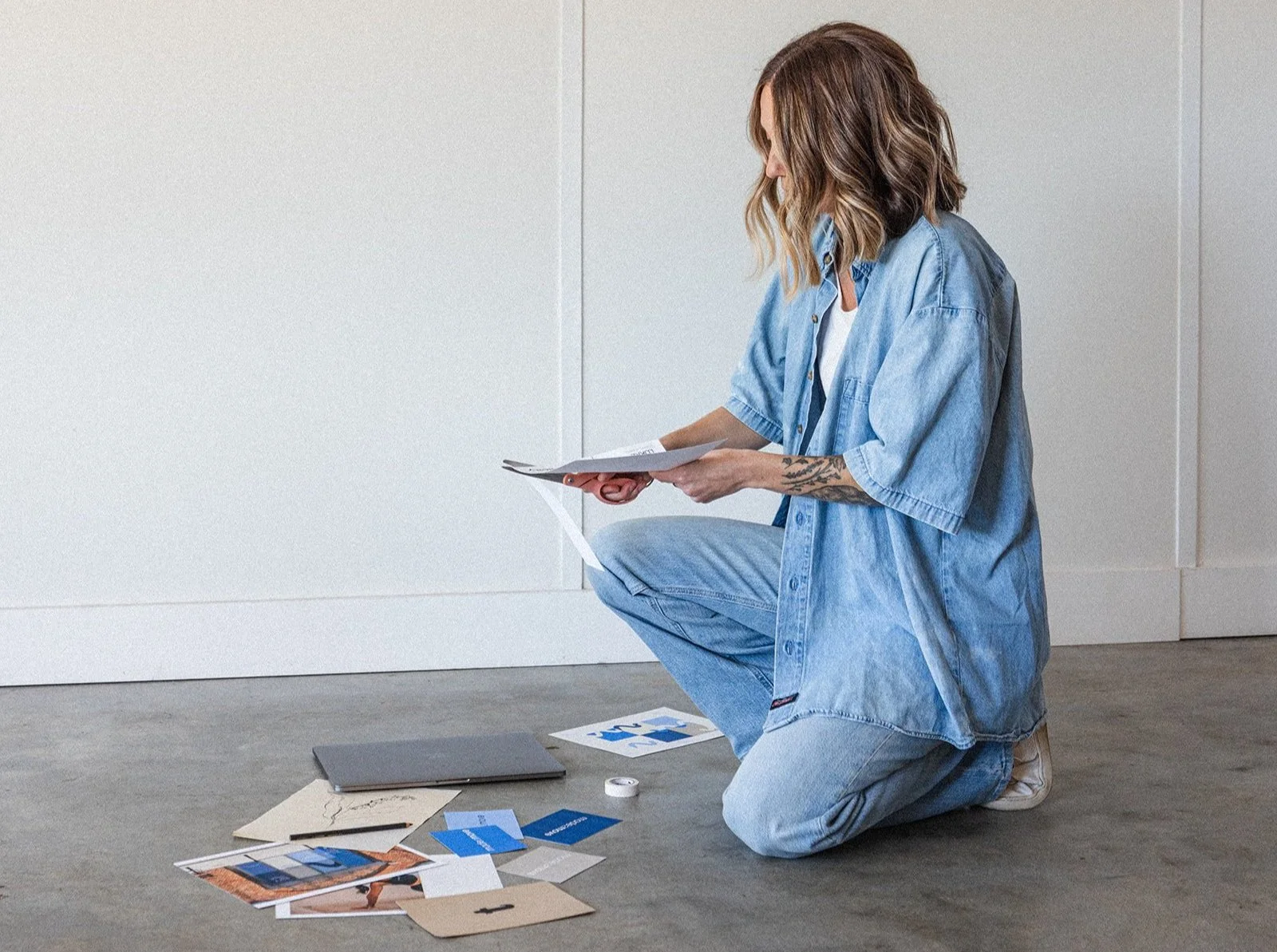 A woman with wavy brown hair and tattoos on her arm is kneeling on the floor, looking at papers she is holding. She is wearing an oversized light blue denim shirt and jeans, with a white shirt underneath. There are various papers, color swatches, and a laptop on the floor in front of her, against a white wall.