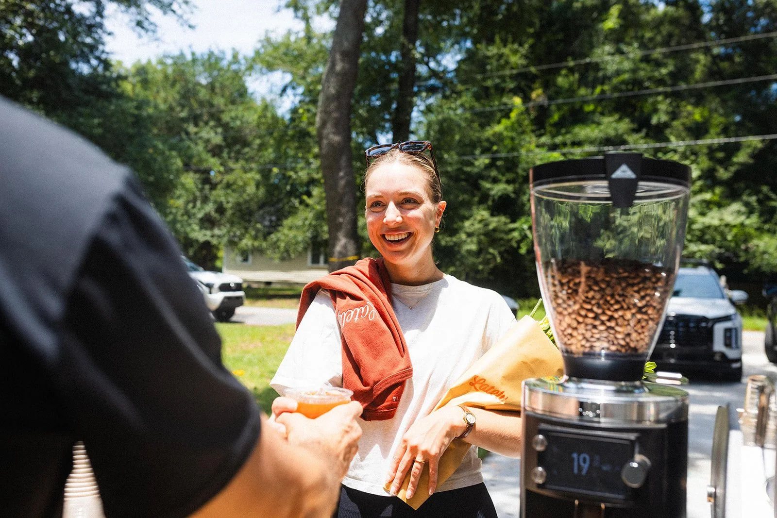 A smiling woman with sunglasses on her head, holding a paper bag, standing outdoors near a coffee grinder filled with coffee beans, with a person handing her a small cup.