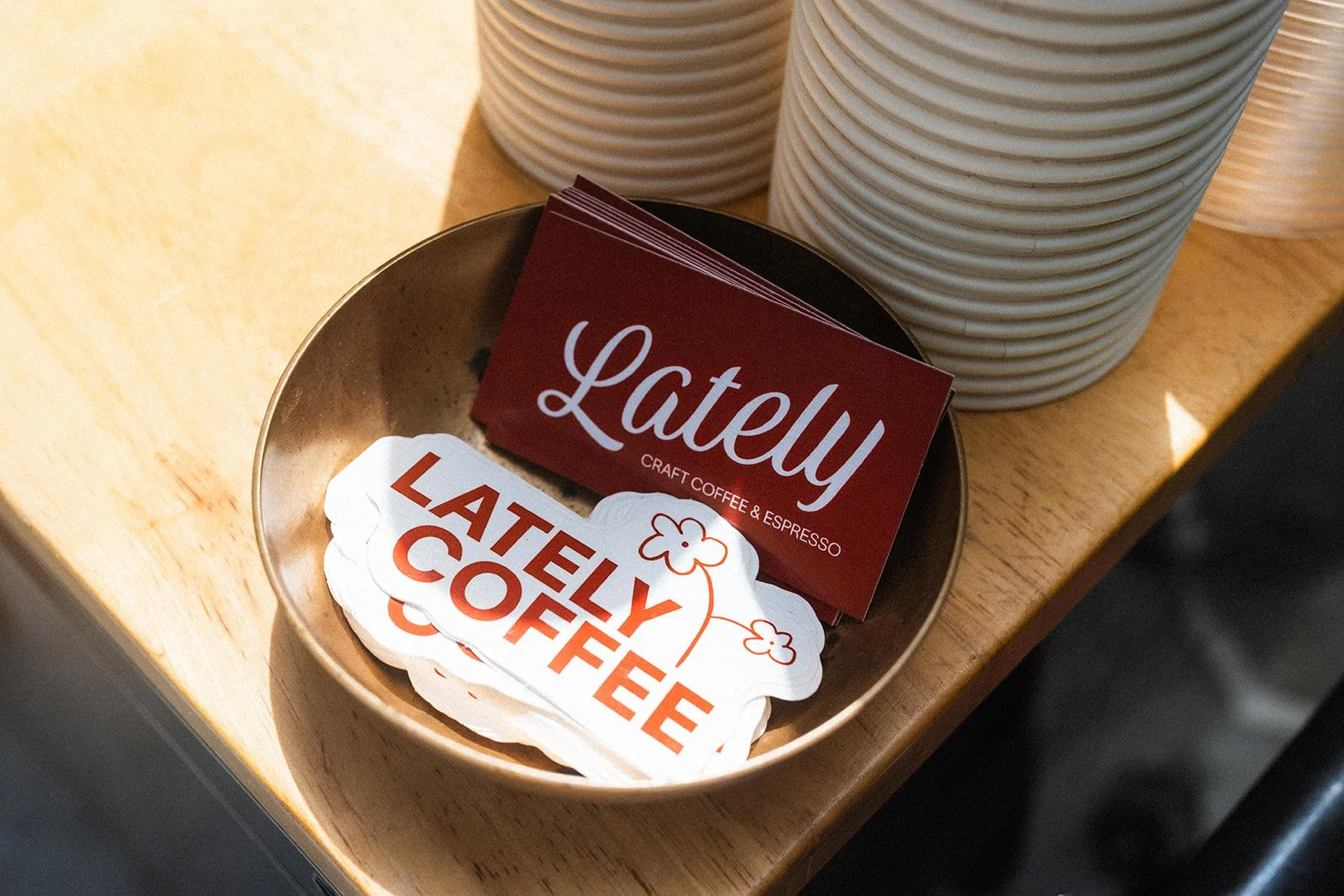 A metal bowl on a wooden table holding red business cards with 'Lately' written on them, and paper coasters that say 'Lately Coffee' with an illustration of a flower, with white cups in the background.