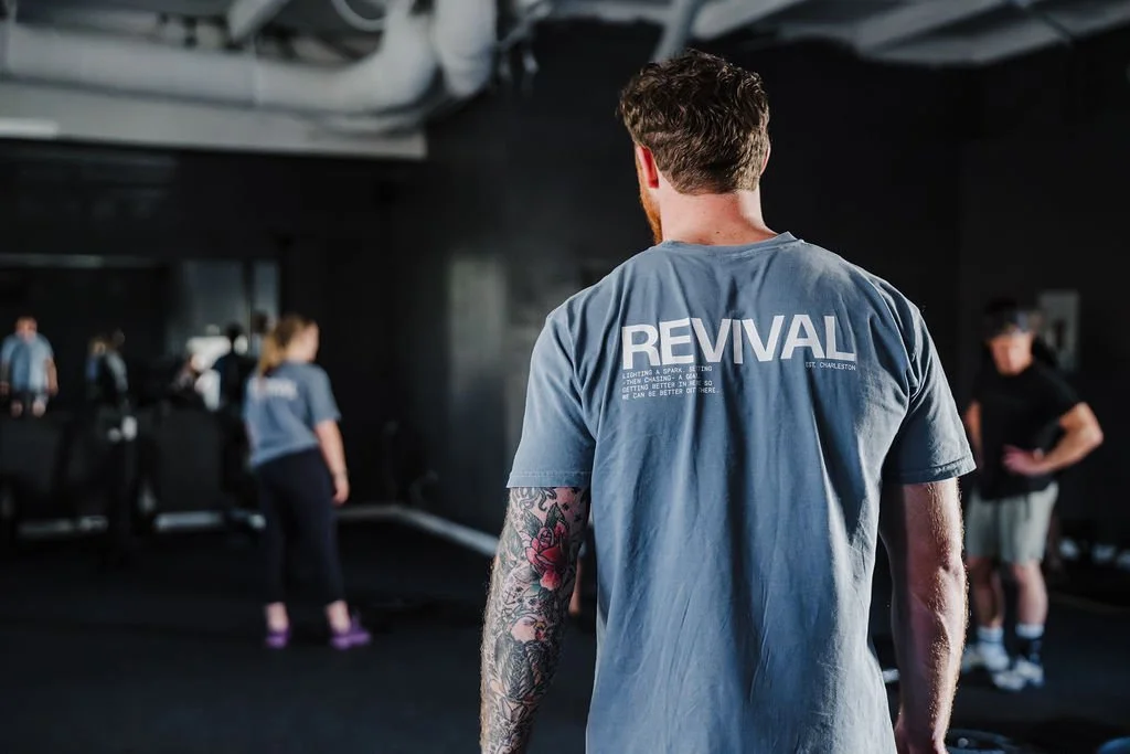 Man with tattooed arm in a blue "REVIVAL" T-shirt standing in a gym with other people exercising in the background.