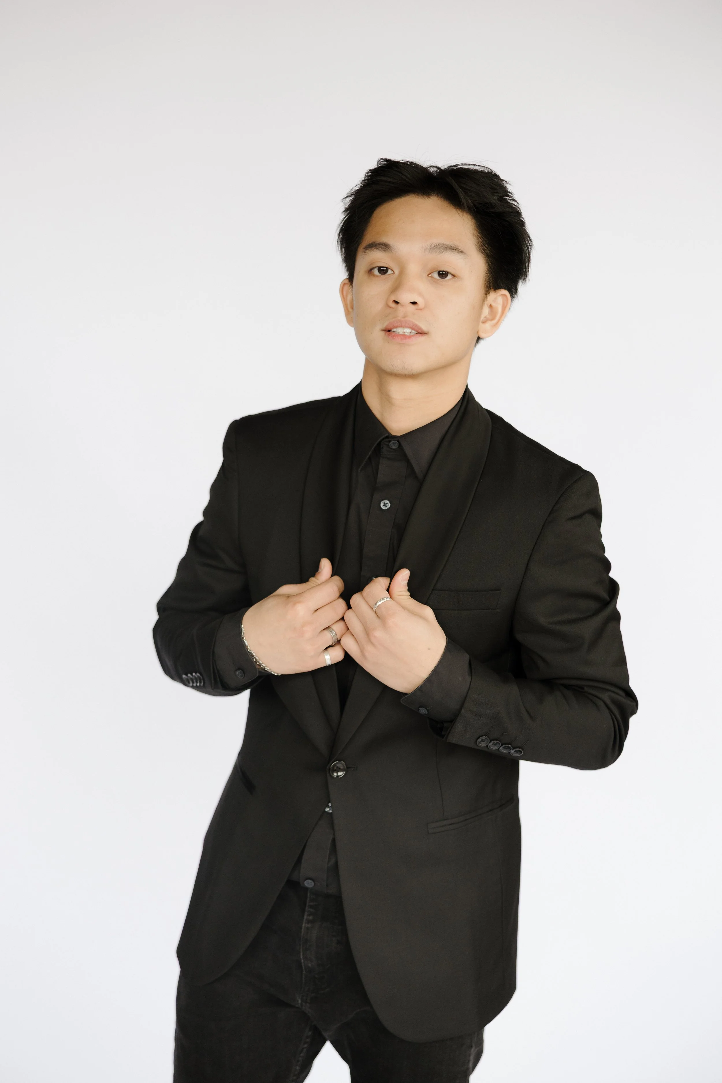 A young man with black hair wearing a black suit and black shirt, standing against a white background.