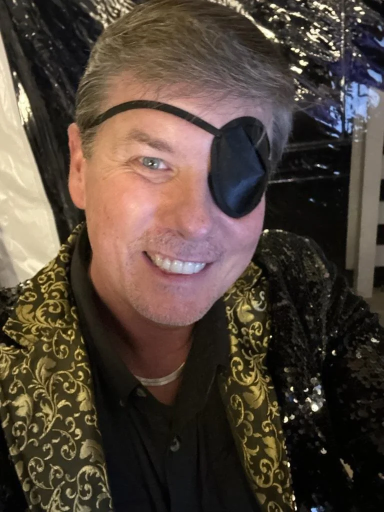 A smiling man with gray hair wearing a black eye patch over one eye, a black shirt, and a jacket with gold ornate patterns and sequins, taking a selfie in front of a shiny black background.
