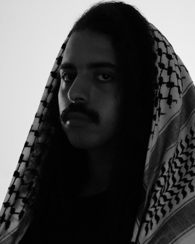 Black-and-white portrait of a mustached person with long hair draped in a patterned keffiyeh scarf