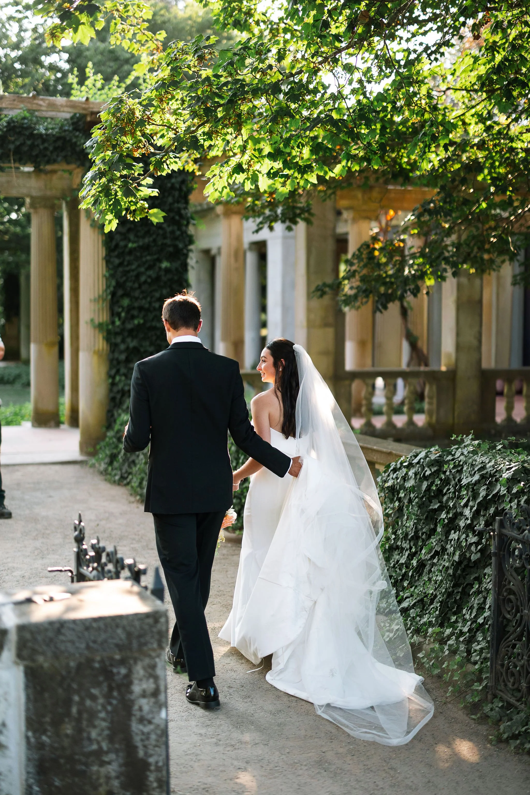 groom-carrying-brides-train-through-garden-eolia-mansion-waterford-conneticut-editorial-wedding