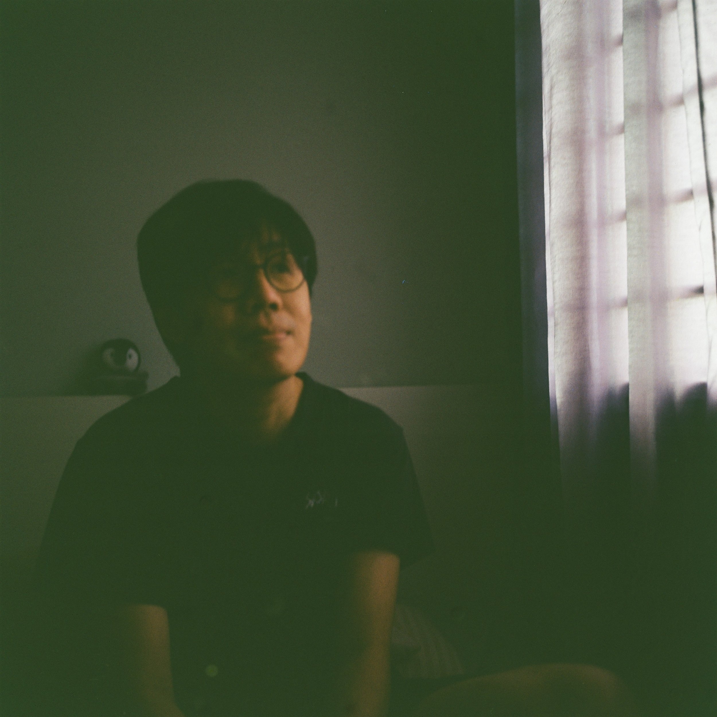 Person with glasses sitting in a dimly lit room near a window with curtains.