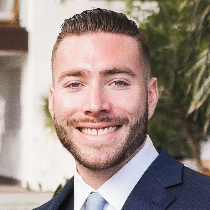 A smiling man with short hair and a trimmed beard, wearing a blue suit and tie, standing outdoors with greenery in the background.