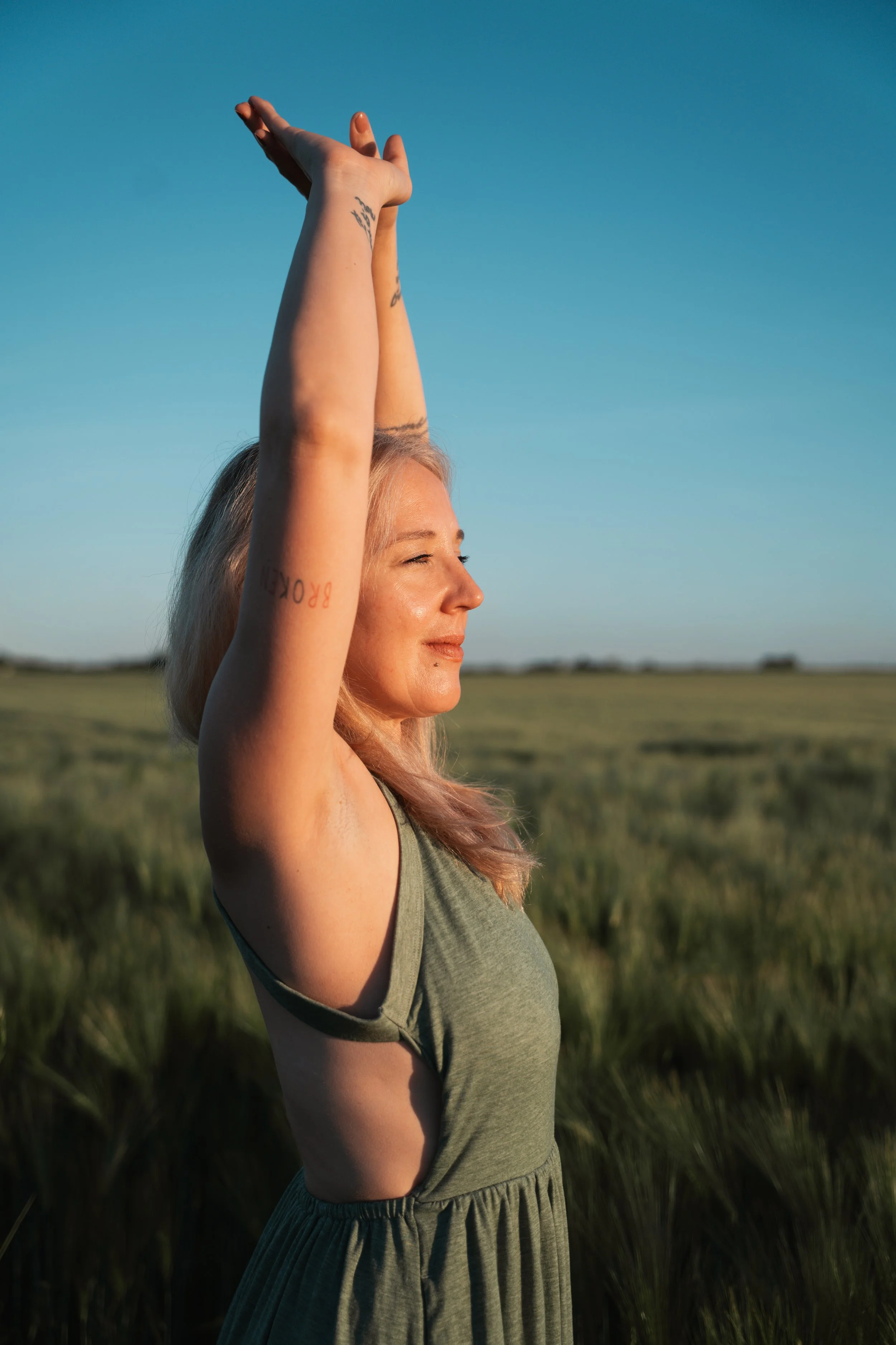 Eine junge Frau mit blondem Haar steht in einem offenen Feld bei Sonnenuntergang, hebt die Arme über den Kopf und macht eine Yoga-Pose, während sie die Augen schließt.