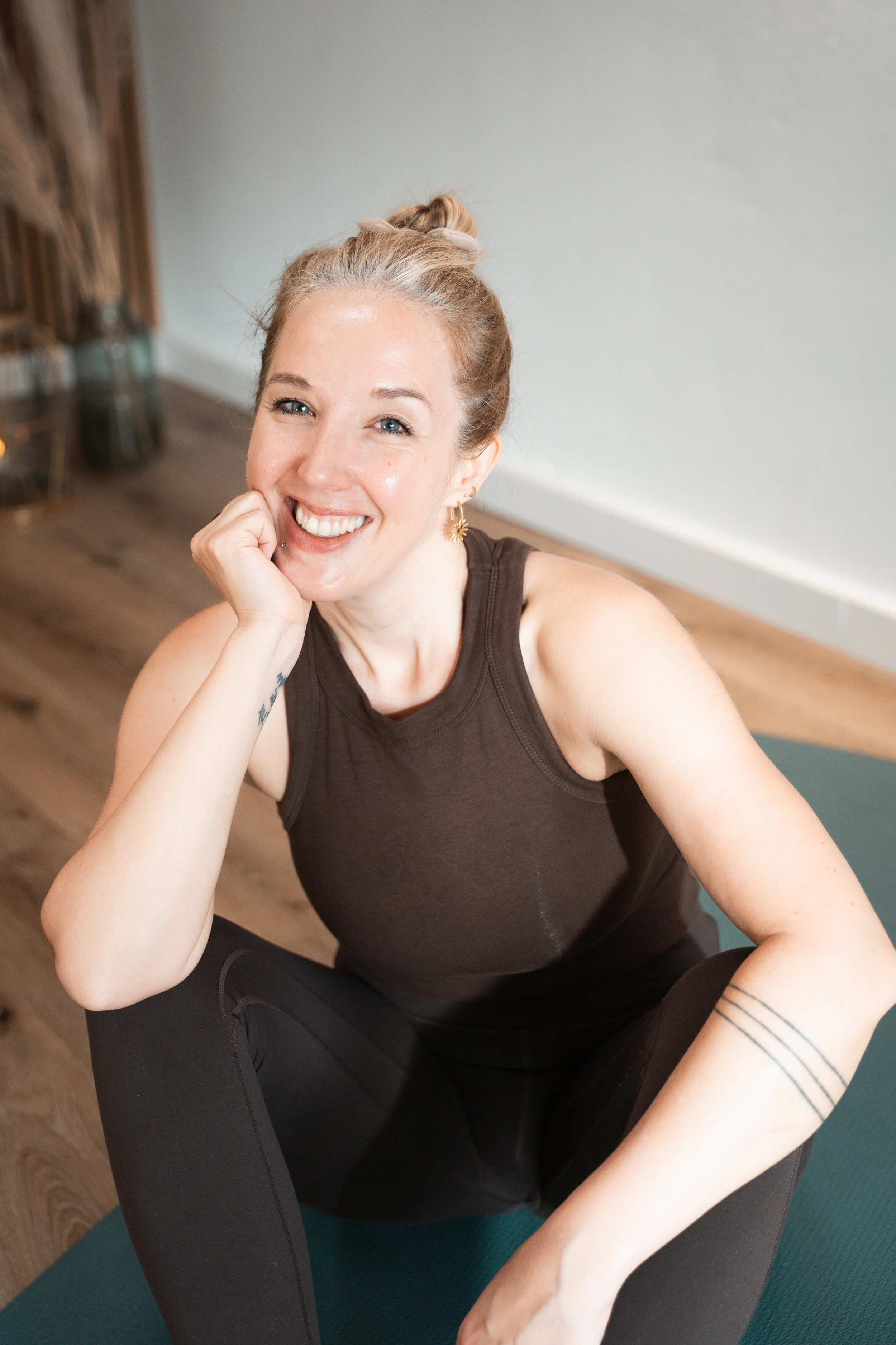 Eine lachende Frau sitzt im Yogaraum auf einer Yogamatte, heben sie den Ellenbogen und trägt einen braunen Tanktop. Im Hintergrund sind Holzbalken und eine weiße Wand.