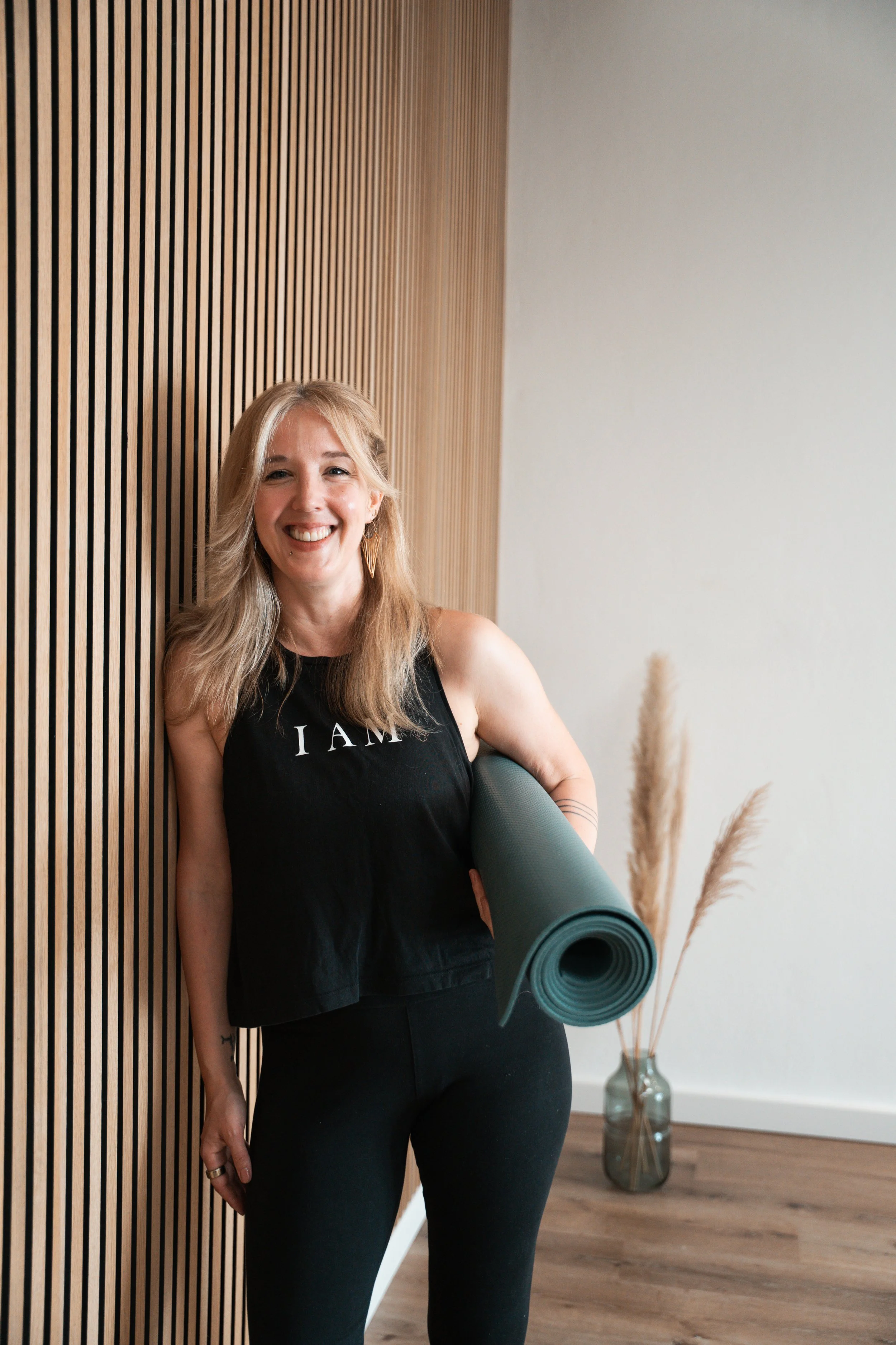 Eine lachende Frau mit langen blonden Haaren trägt schwarze Kleidung und hält eine Yogamatte in einem Raum mit Holzwand und Dekorationen. Sie steht neben einer Vase mit Trockenpflanzen.