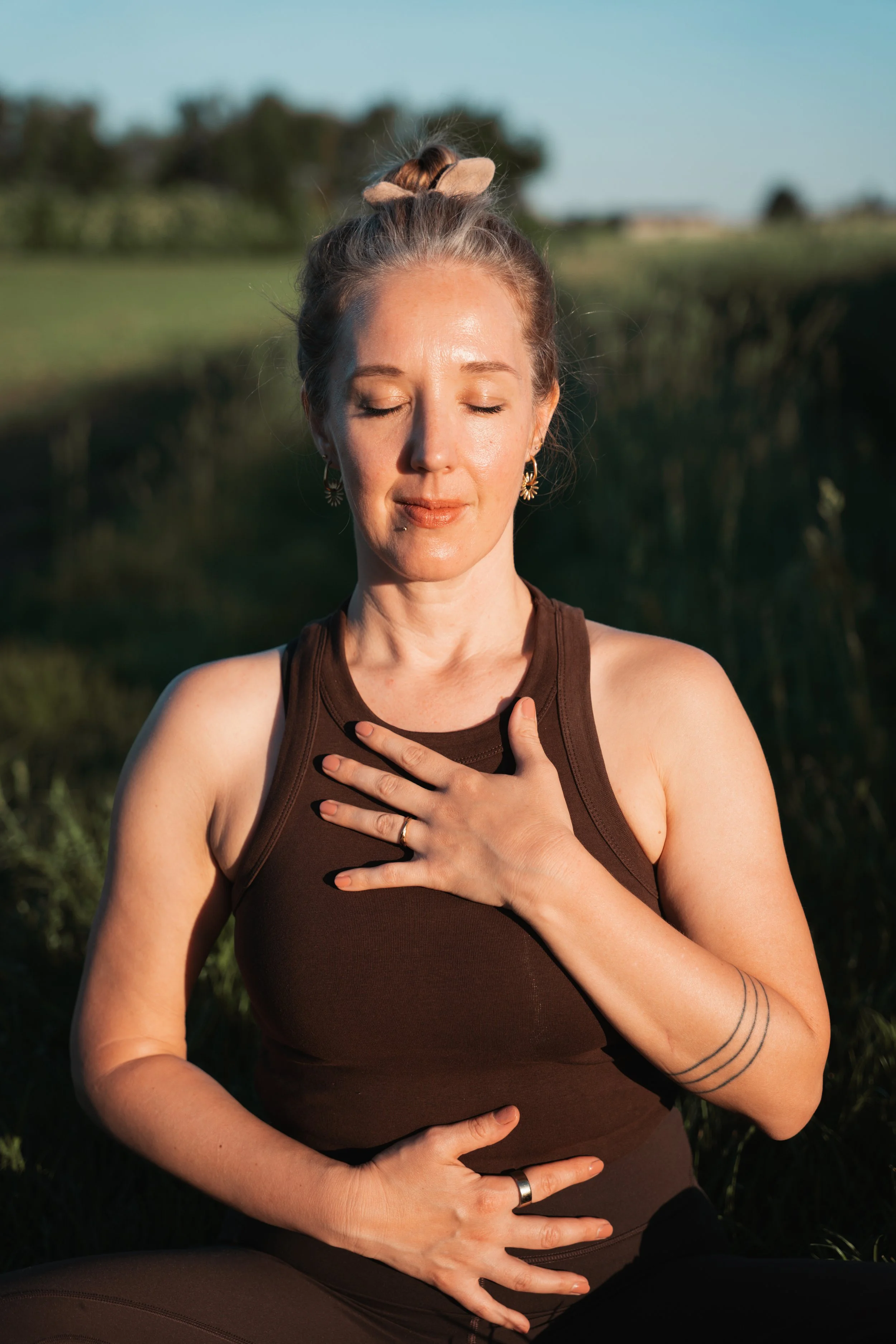 Eine Frau übt Yoga im Freien, sitzt im Schneidersitz mit geschlossenen Augen, Hand auf der Brust, bei Sonnenuntergang in einer natürlichen Umgebung.