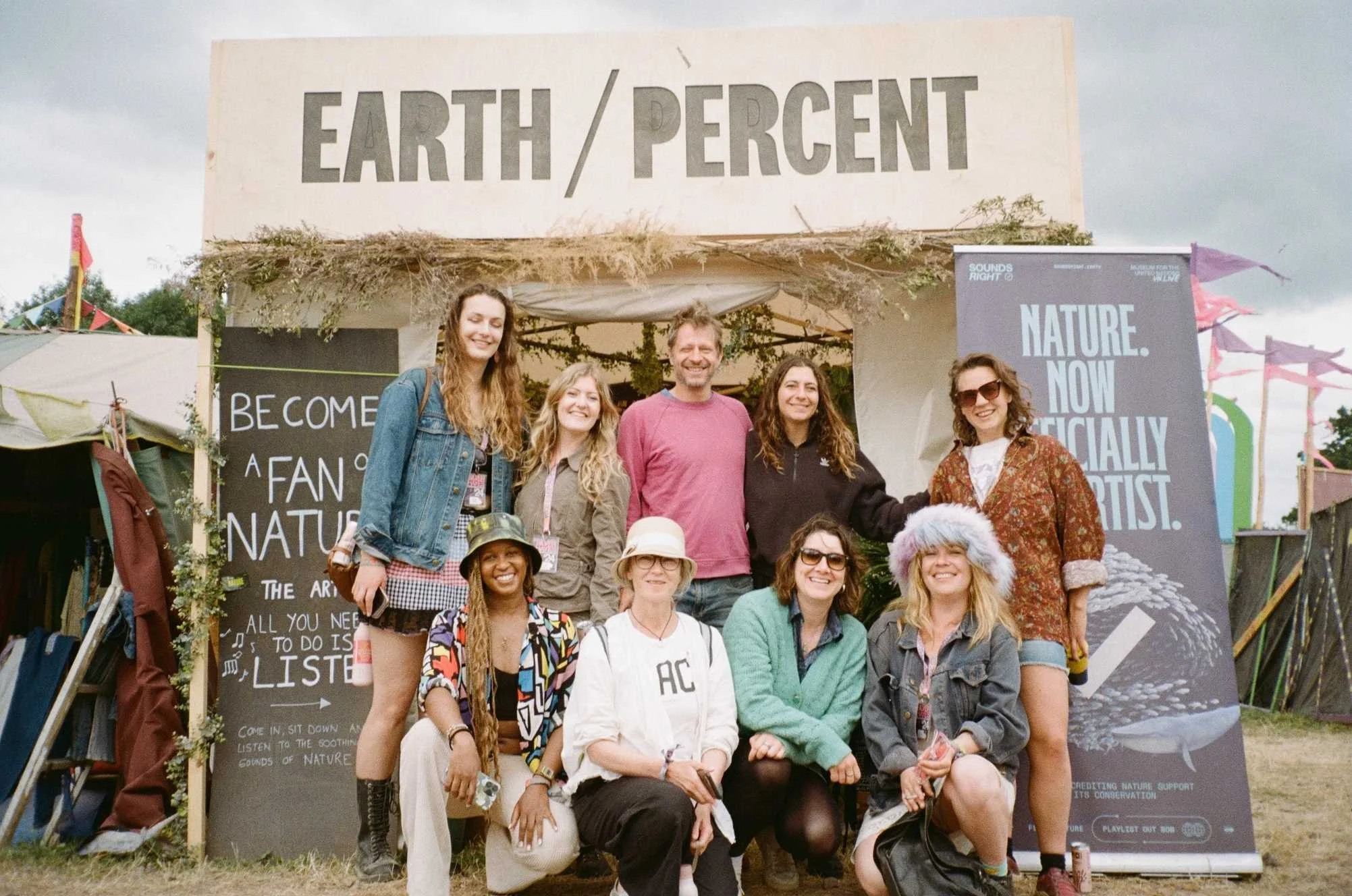 Group photo of nine people smiling at the camera and standing at an outdoor festival in front of a sign Earth / Percent.