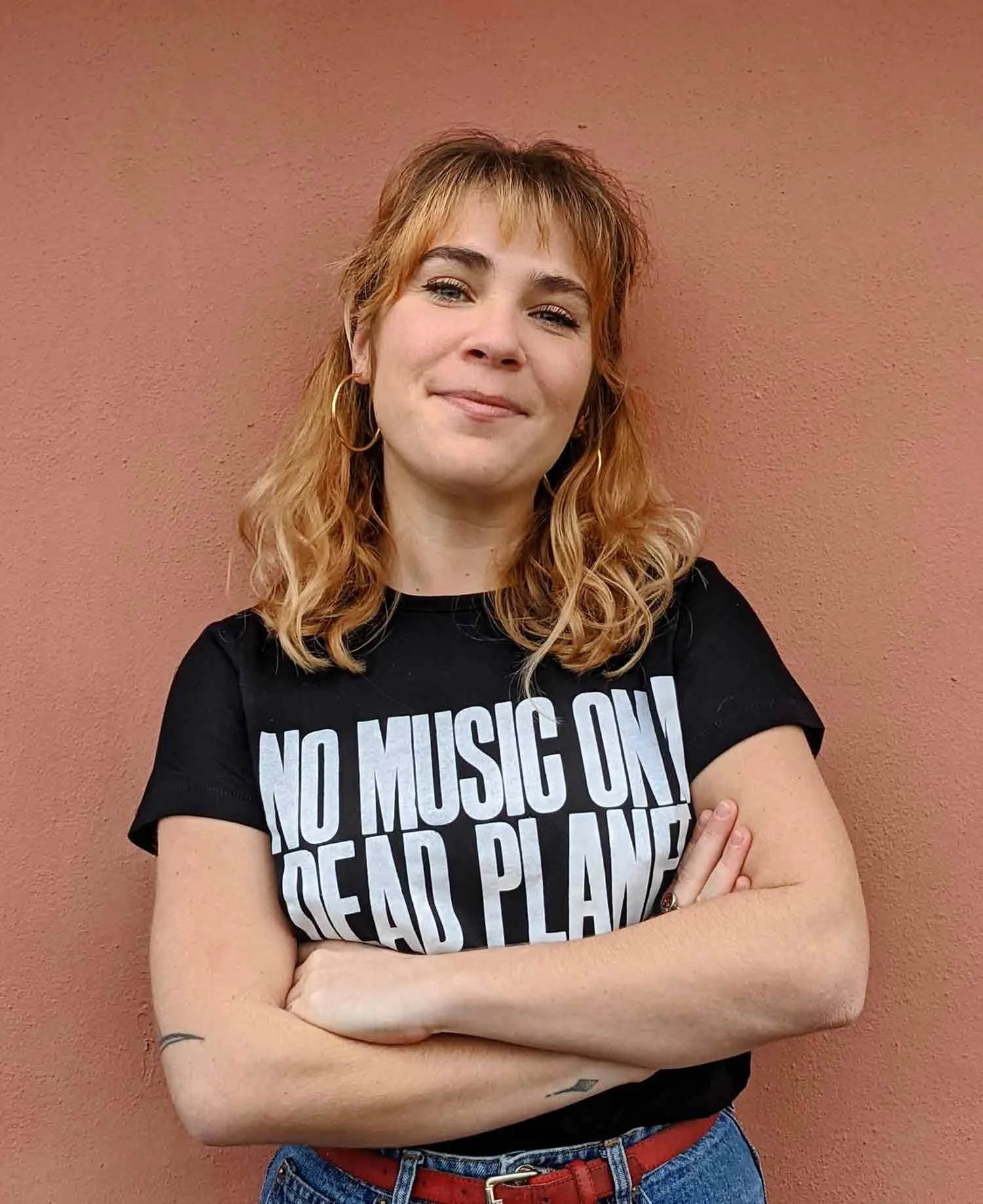 Women standing in front of wall with arms crossed and wearing a t-shirt with text no music on a dead planet