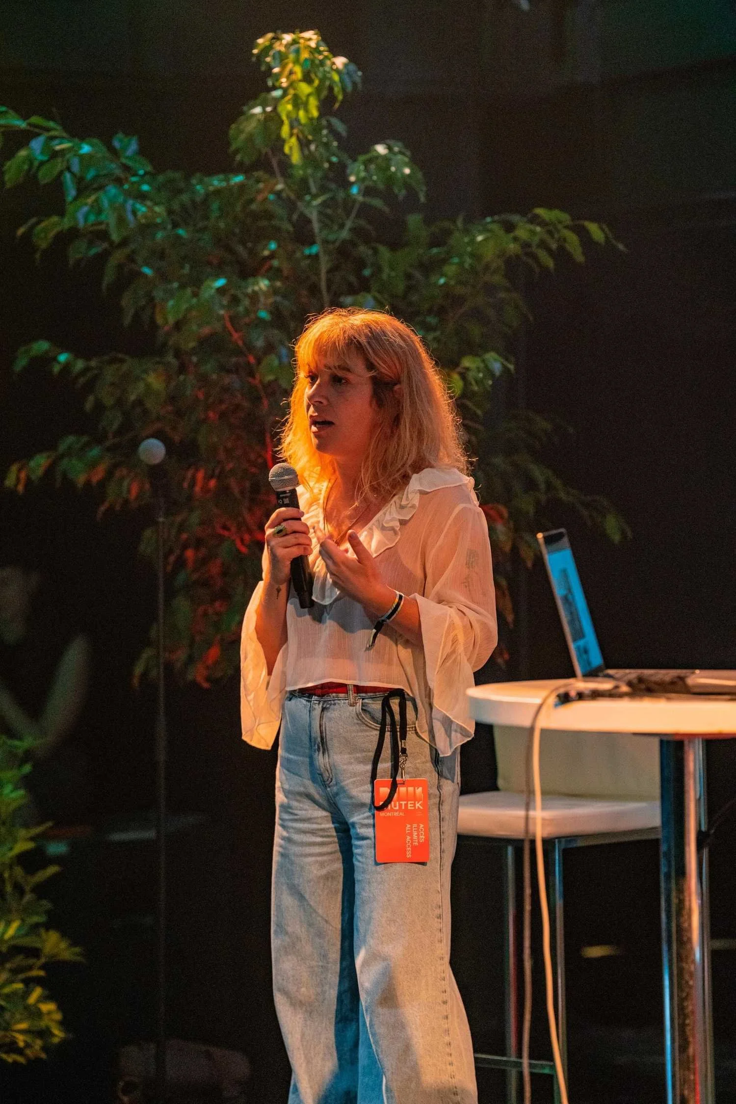 Pauline Bourdon standing on stage with a microphone in hand giving a talk wearing a white ruffled blouse and light blue jeans.