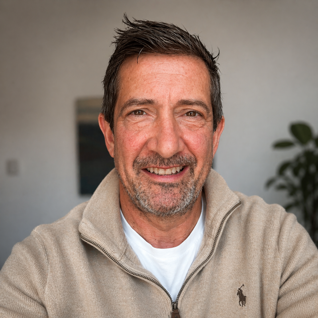 A smiling middle-aged man with dark hair and facial hair, wearing a beige quarter-zip sweater with a Polo logo, sitting indoors with a blurred background that includes a small plant and a painting or photograph on the wall.