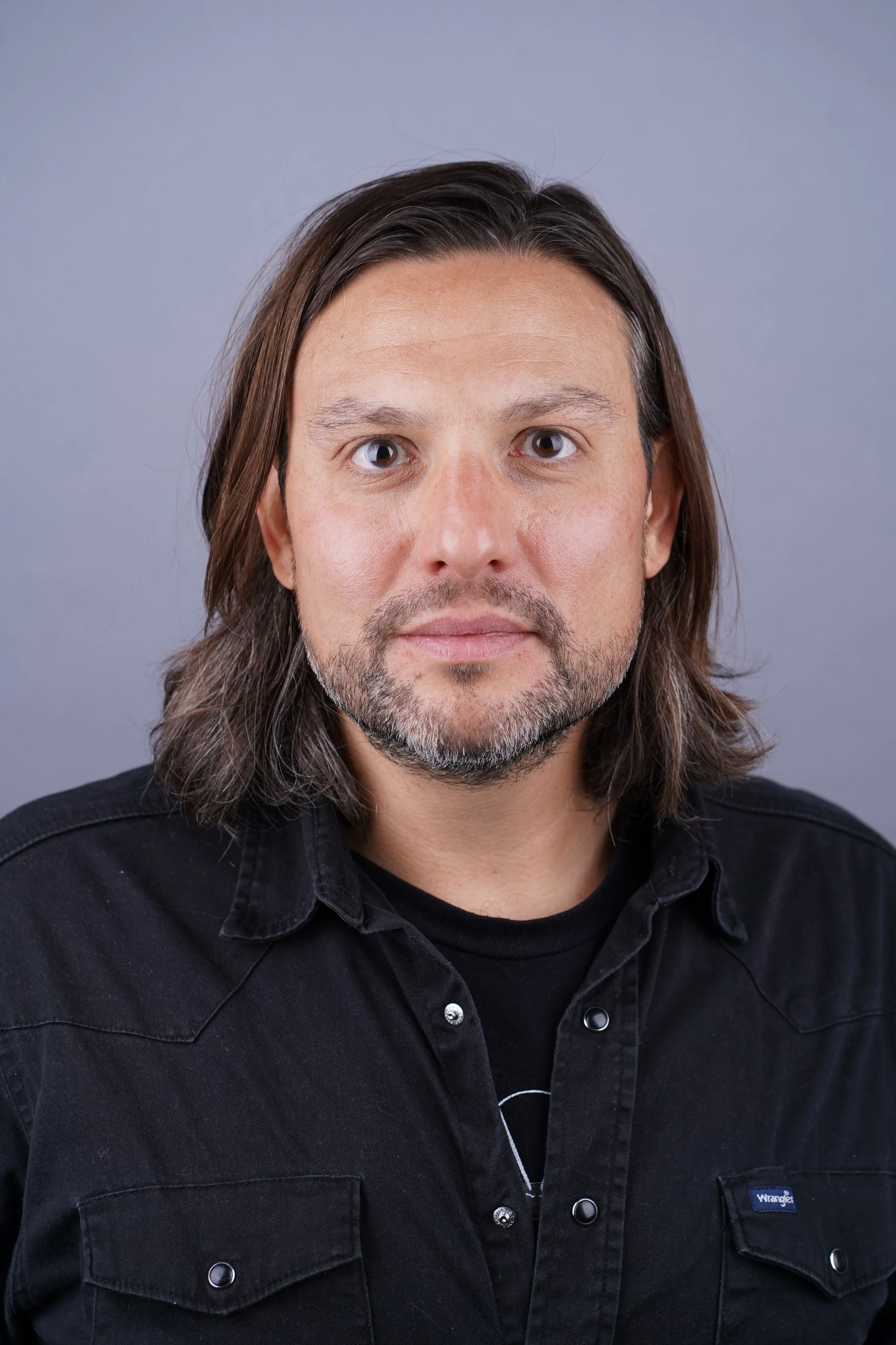 A middle-aged man with long brown hair, a beard, and hazel eyes, wearing a black Wrangler shirt, looking directly at the camera against a plain gray background.