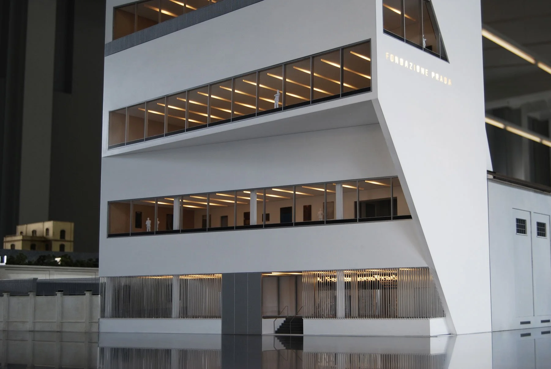 Moderna struttura architettonica bianca con grandi finestre, illuminata dall'interno, chiamata Fondazione Prada, situata vicino a un specchio d'acqua che riflette l'edificio.