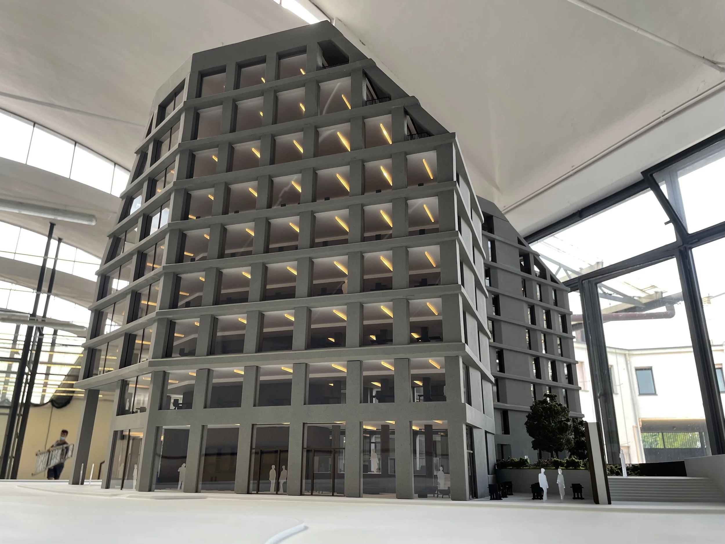 Modello architettonico di un edificio moderno a più piani, situato all'interno di un'area vetrata con grandi finestre e luci interne, con alcune piante e persone che si muovono nell'ambiente.