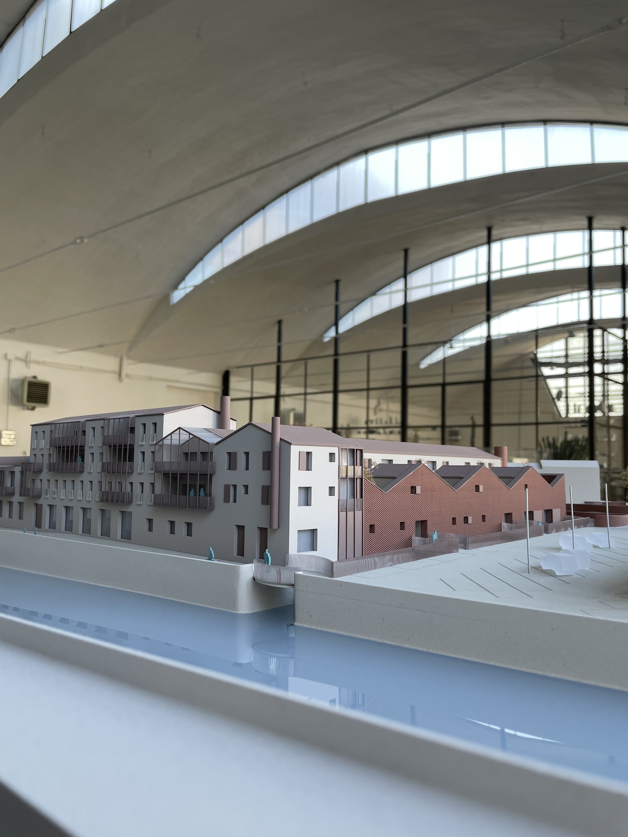 Modello di un complesso residenziale con edifici a più piani e tetti inclinati, situato su una piattaforma circondata da un muro di recinzione, con un'area di parcheggio e un ruscello artificiale in primo piano.