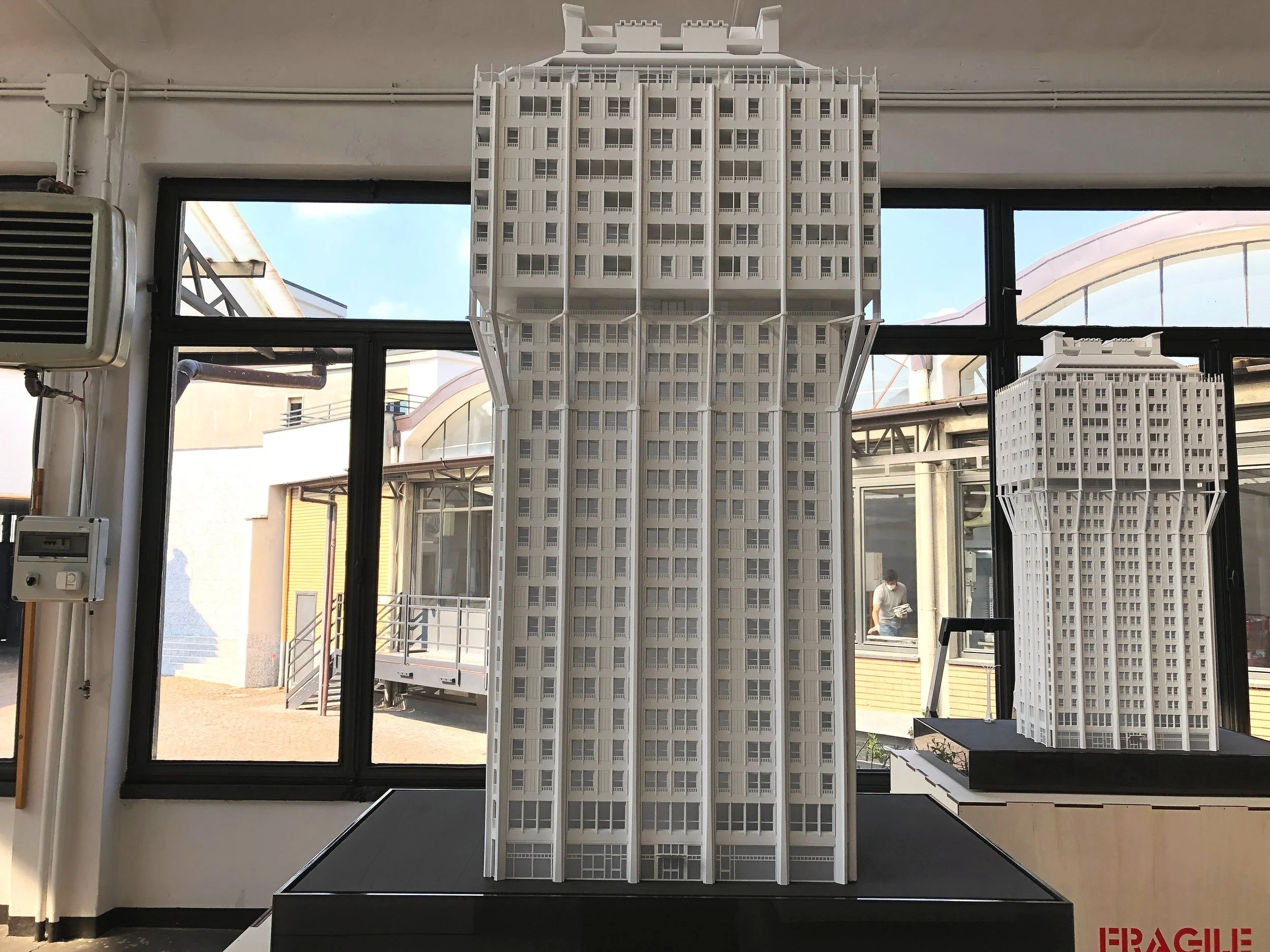 Modello architettonico di un edificio alto con molti piani, realizzato in materiali chiari, posizionato di fronte a una grande finestra con vista su edifici circostanti.