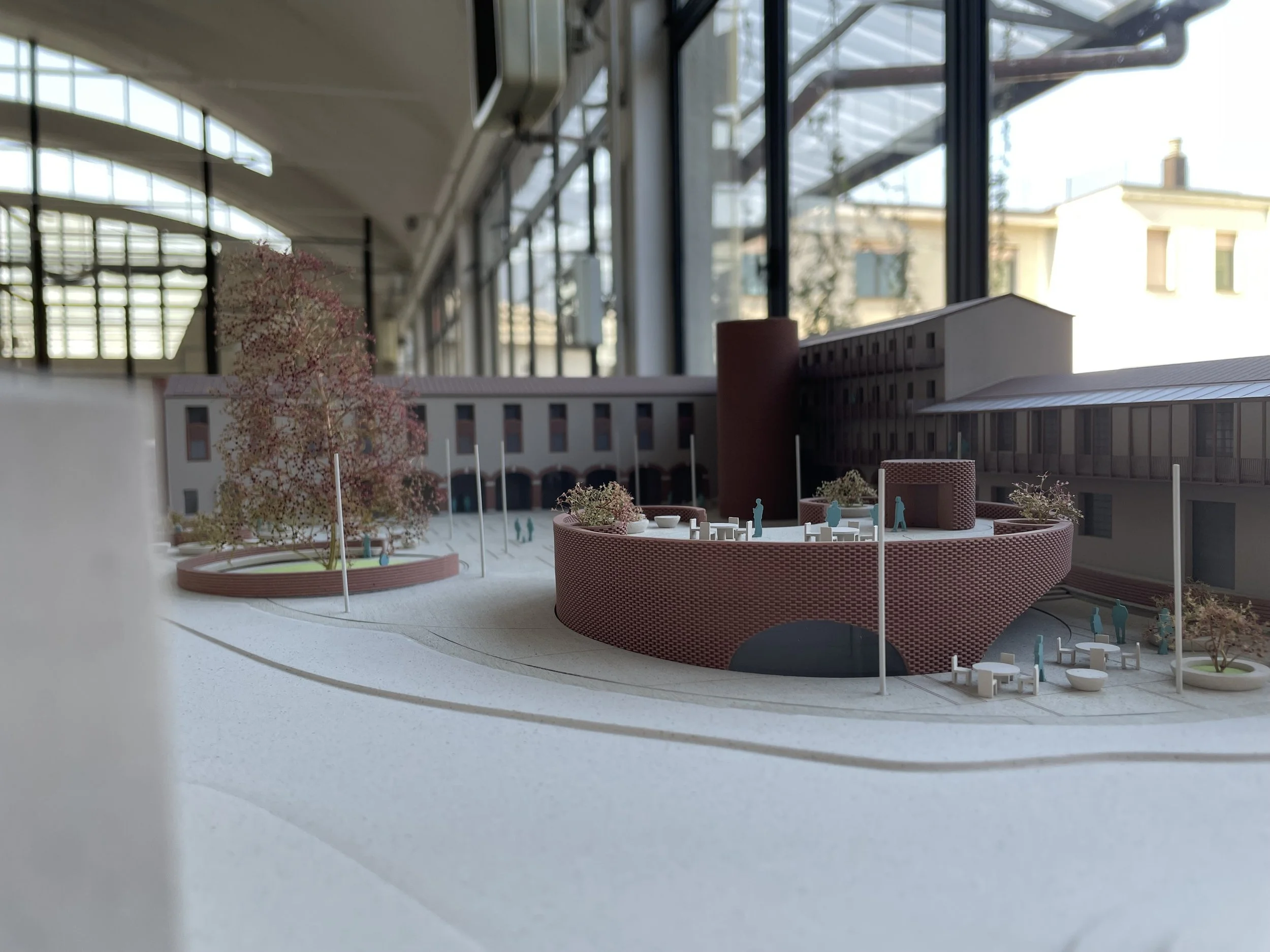 Modello di un'architettura moderna in scala, raffigurante un edificio con cortile interno e elementi di decorazione con alberi e panchine, all'interno di un ambiente con grandi finestre.