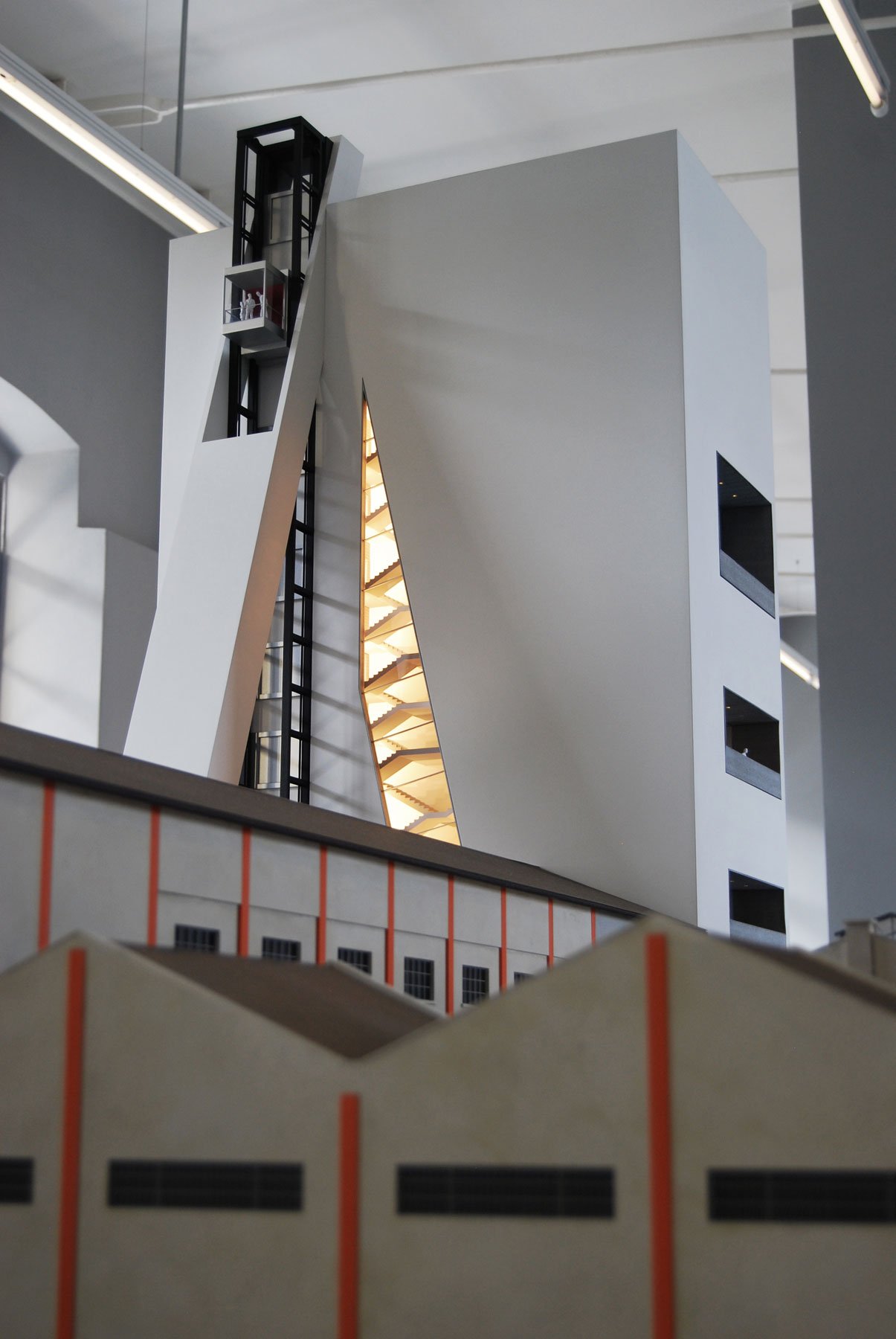 Modello di un edificio moderno con forme geometriche, spazi aperti e illuminazione interna, visibile sul modello in un ambiente espositivo.