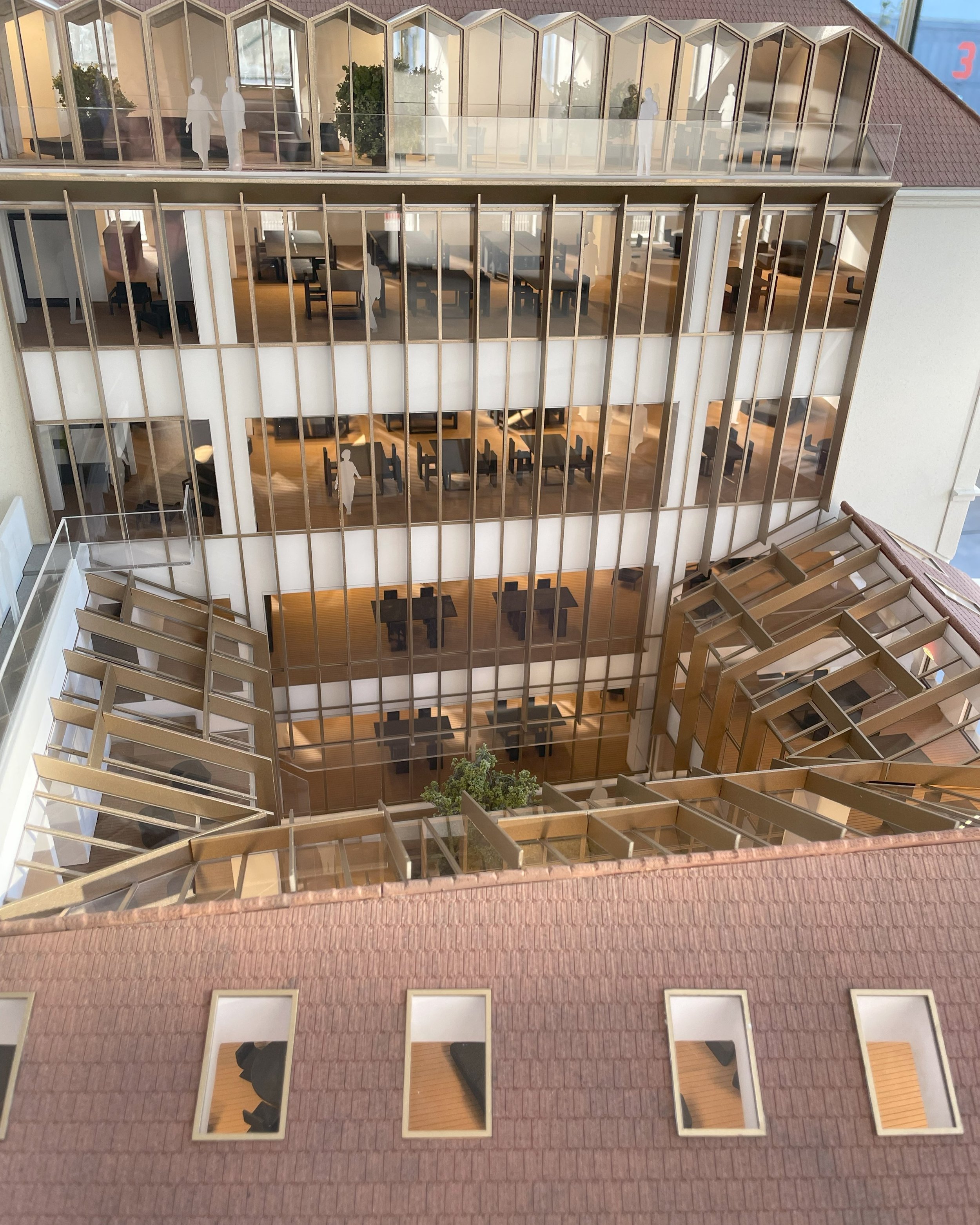 Modello di un edificio con tre piani evidenziati, con pareti in vetro e strutture in legno. Vetrate curvate e muri in mattoni rossi sono visibili, con alcuni spazi interni arredati con tavoli e sedie, e figure umane silhouette.
